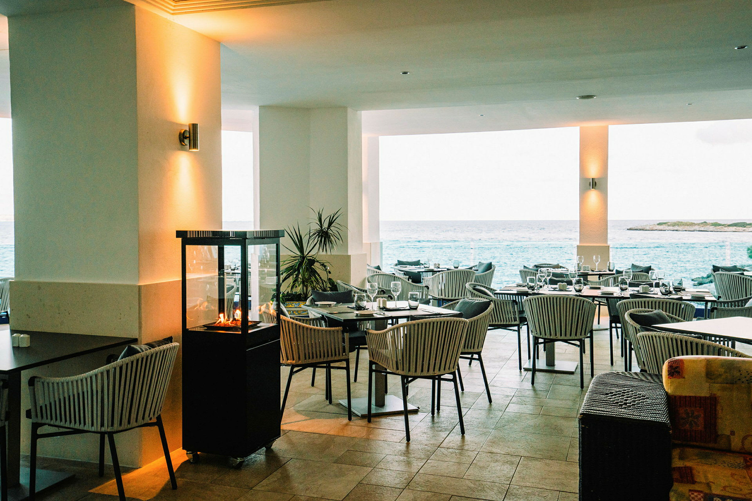 En restaurant med udendørs udsigt over havet, møbleret med borde og stole og dekoreret med en lille pejs. (AI-genereret tekst)