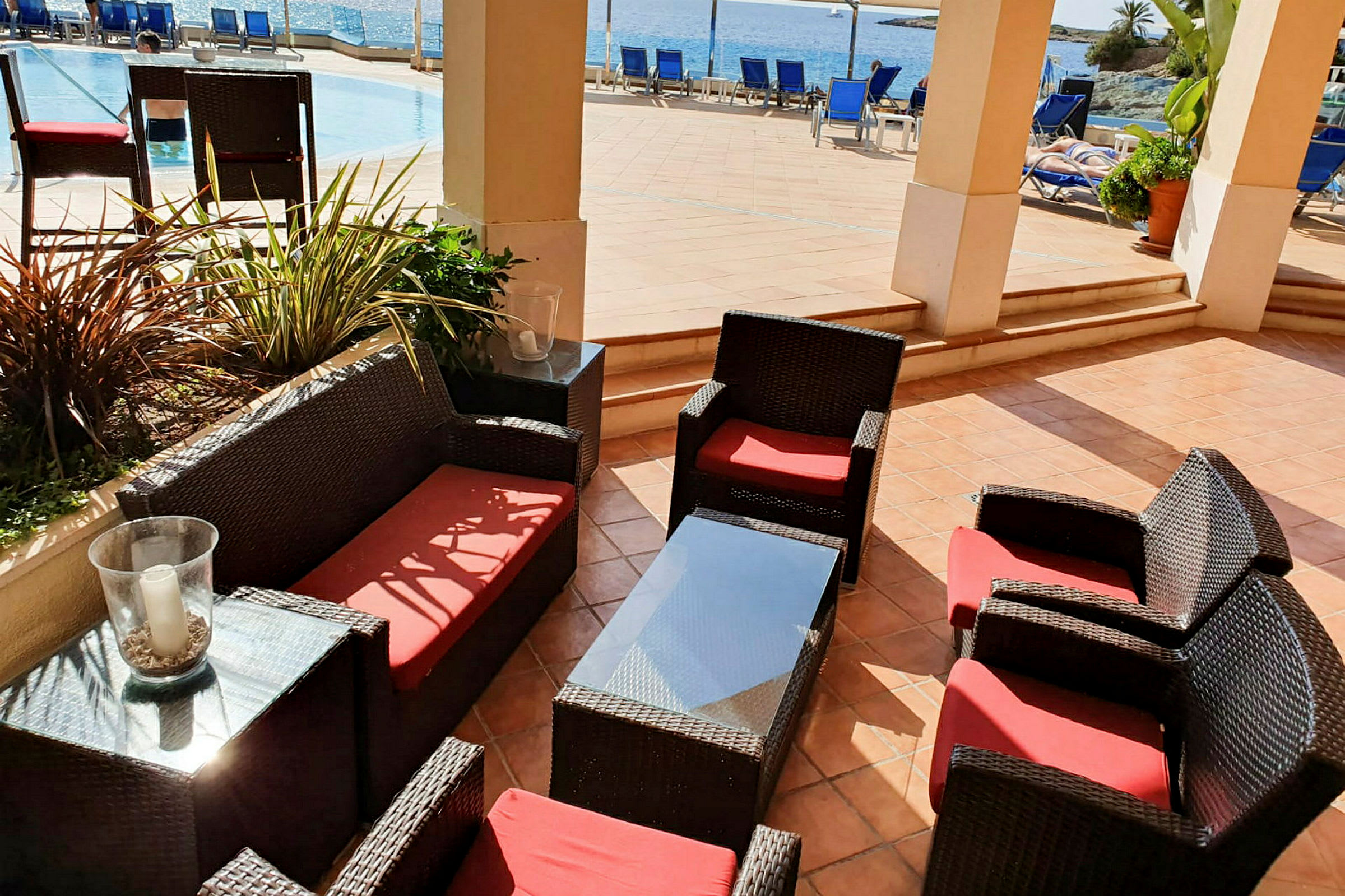 Udendørsmøbler med røde hynder på en terrasse ved pool og hav. (AI-genereret tekst)