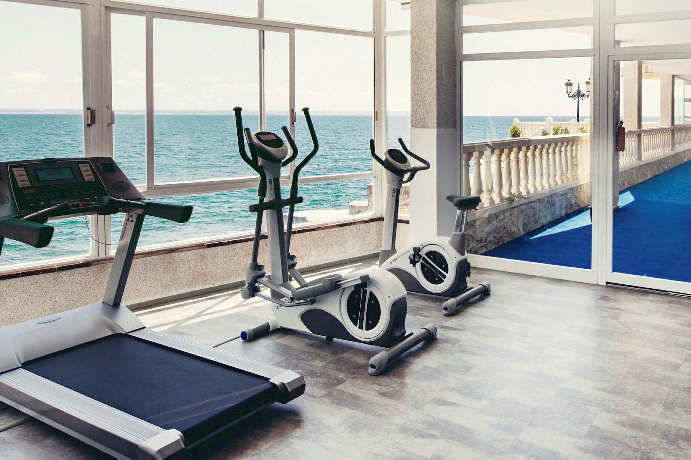 Fitnessrum med træningsudstyr og havudsigt gennem glasvæg. (AI-genereret tekst)