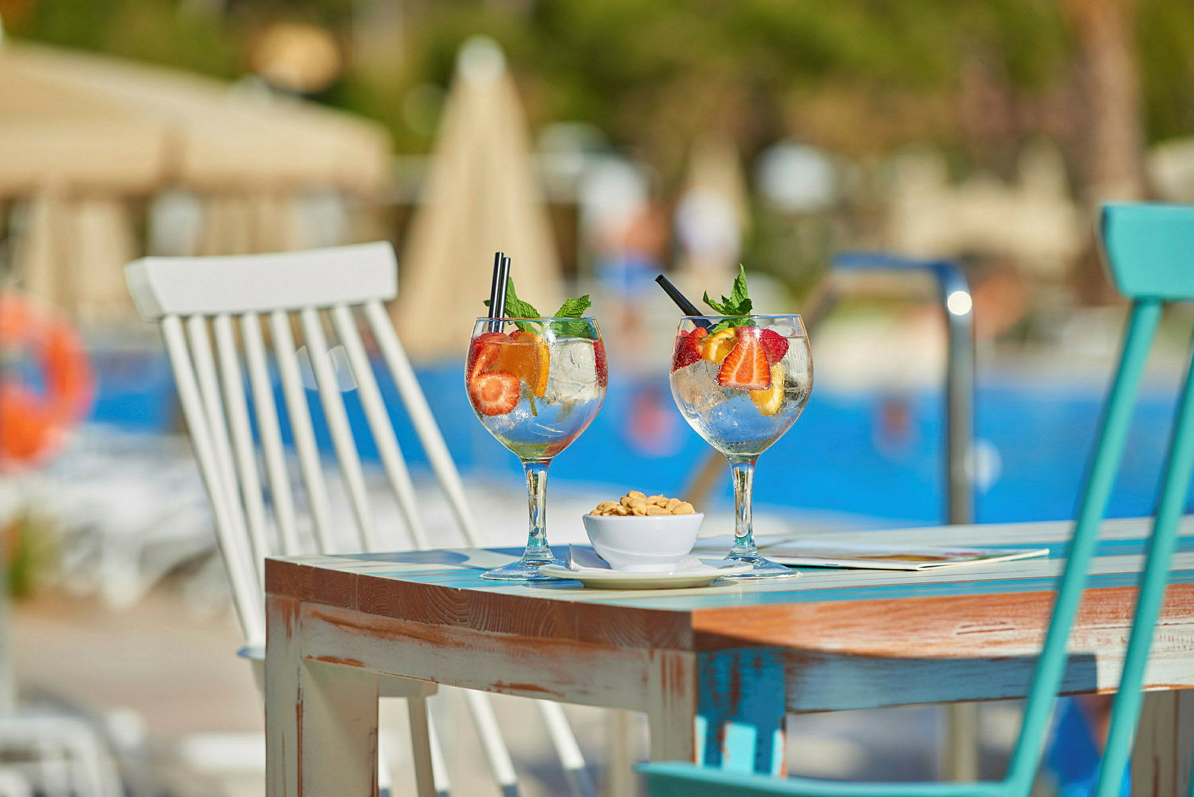 To glas med frugt-cocktails og en skål med snacks på et bord nær en pool. (AI-genereret tekst)