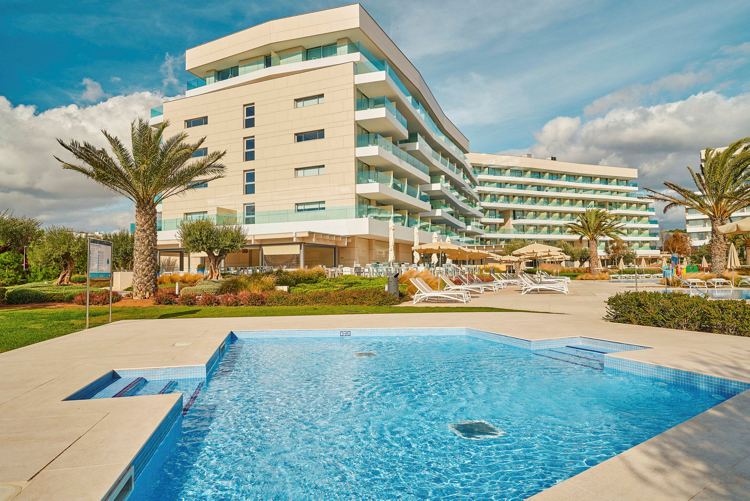 Billede af et hotel med moderne bygninger, en swimmingpool og udendørs loungeområder. (AI-genereret tekst)