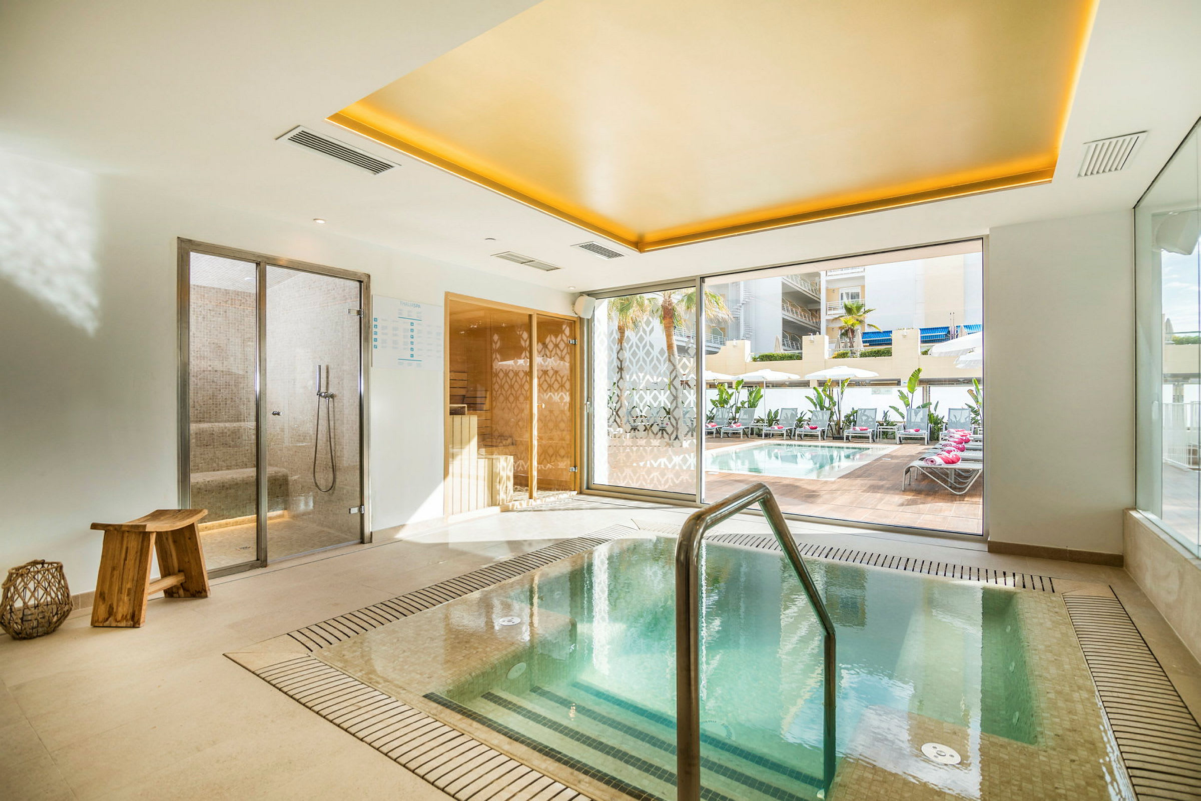 Billede af et indendørs spa med varmt bassin og udsigt til en udendørs pool gennem store vinduer. (AI-genereret tekst)