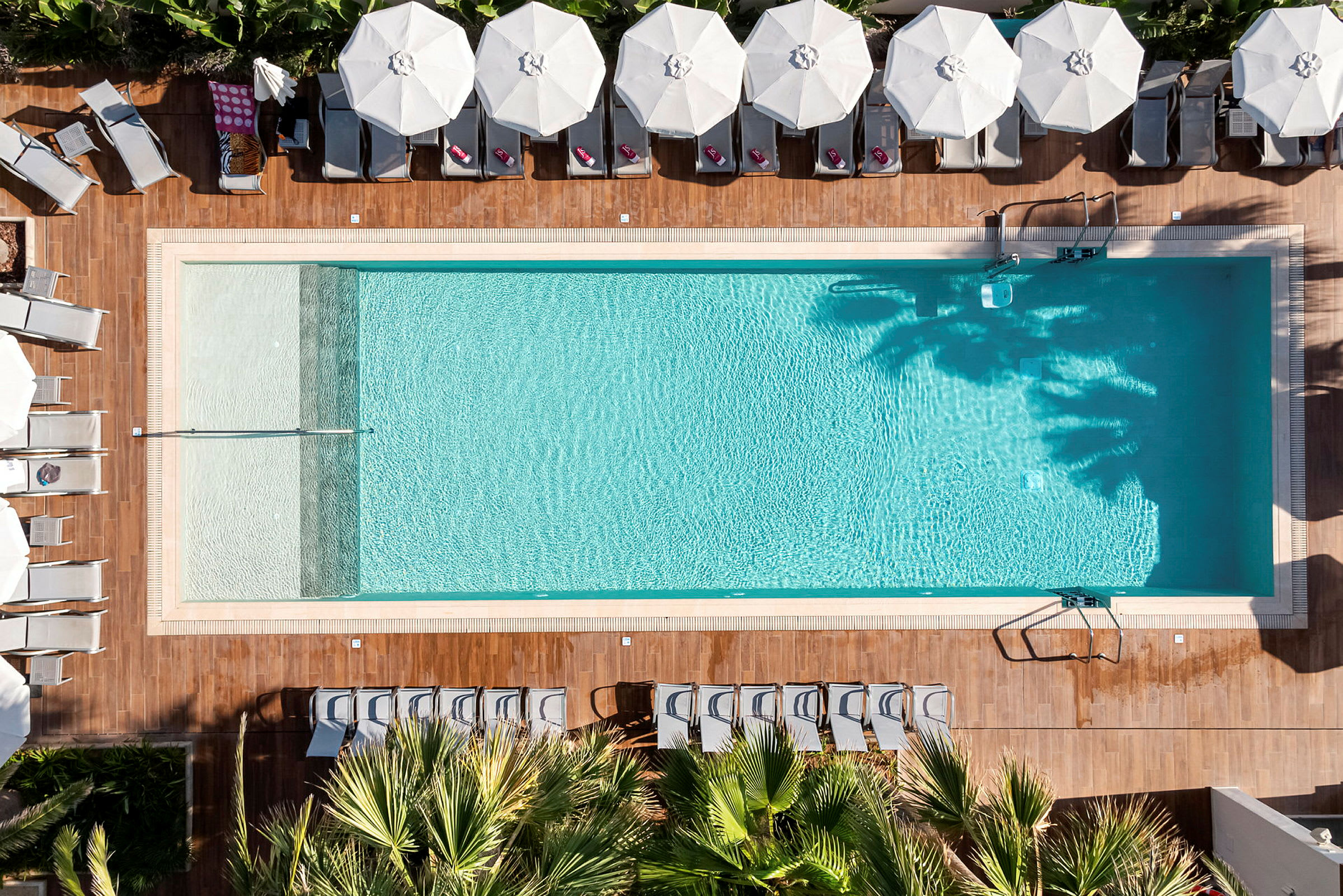 Luftfoto af en rektangulær swimmingpool omgivet af liggestole og parasoller. (AI-genereret tekst)