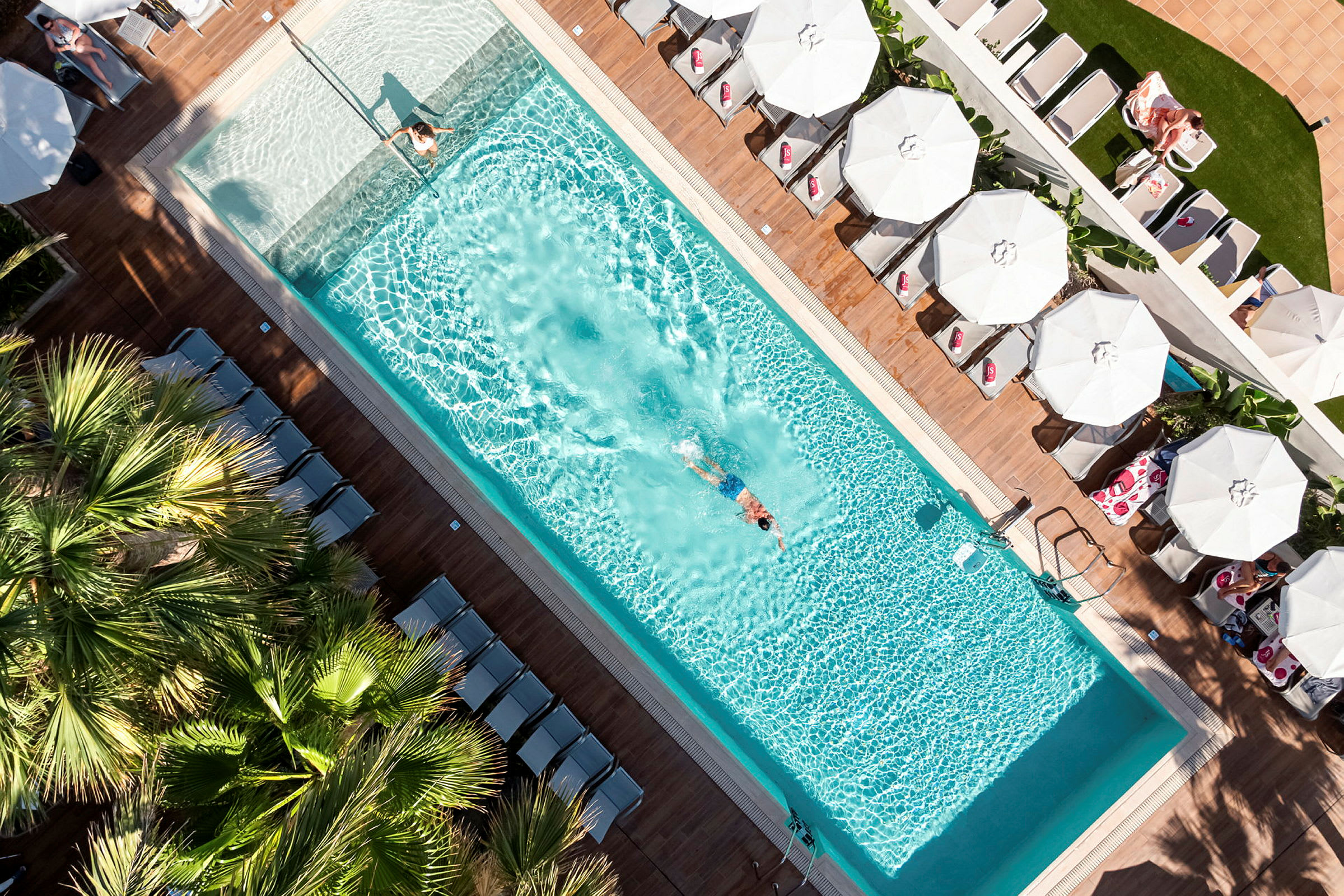 Et fugleperspektiv af en swimmingpool med en svømmende person og liggestole med parasoller omkring. (AI-genereret tekst)