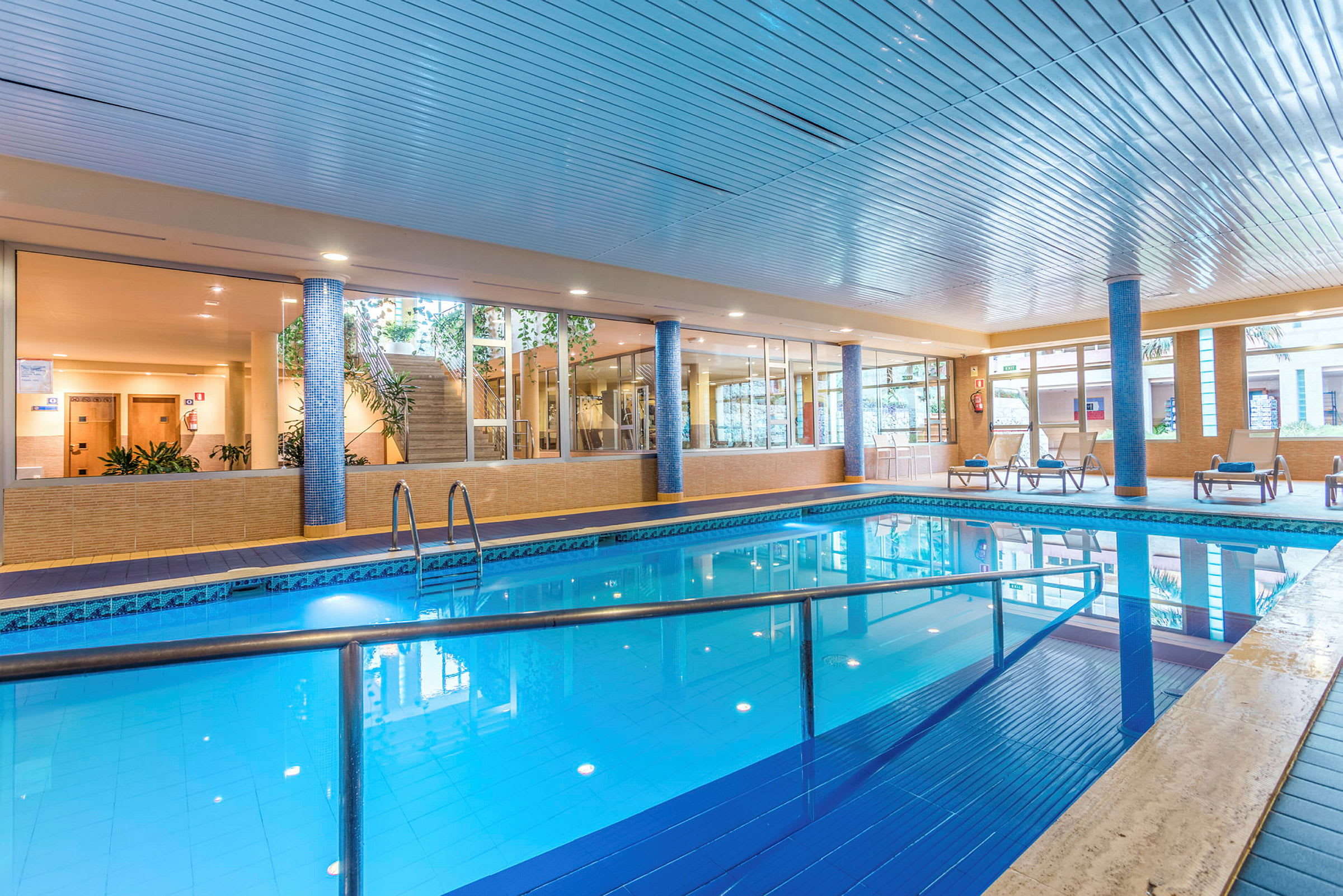 Indendørs pool med liggestole og omkringliggende glasvægge. (AI-genereret tekst)