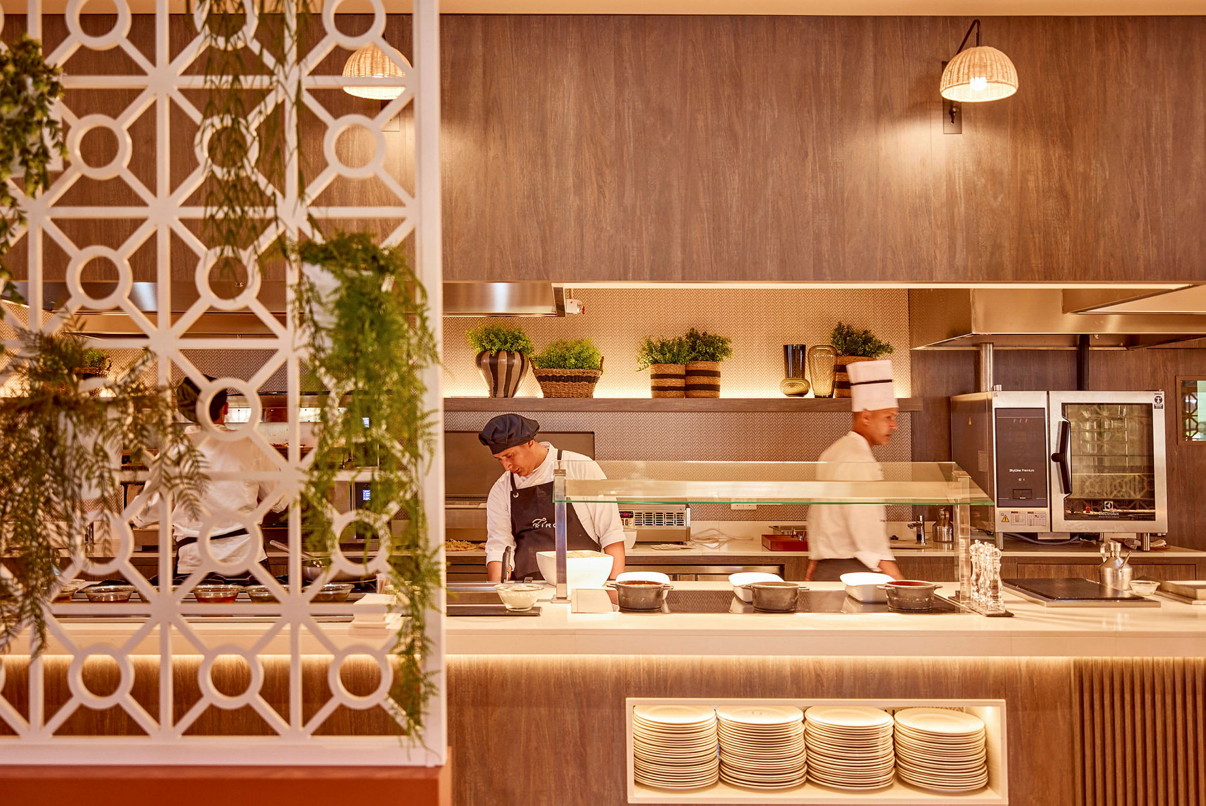 Et restaurantkøkken med kokke i arbejde, en dekorativ væg med hængende planter, tallerkener på hylder og interiør med rustikke detaljer. (AI-genereret tekst)