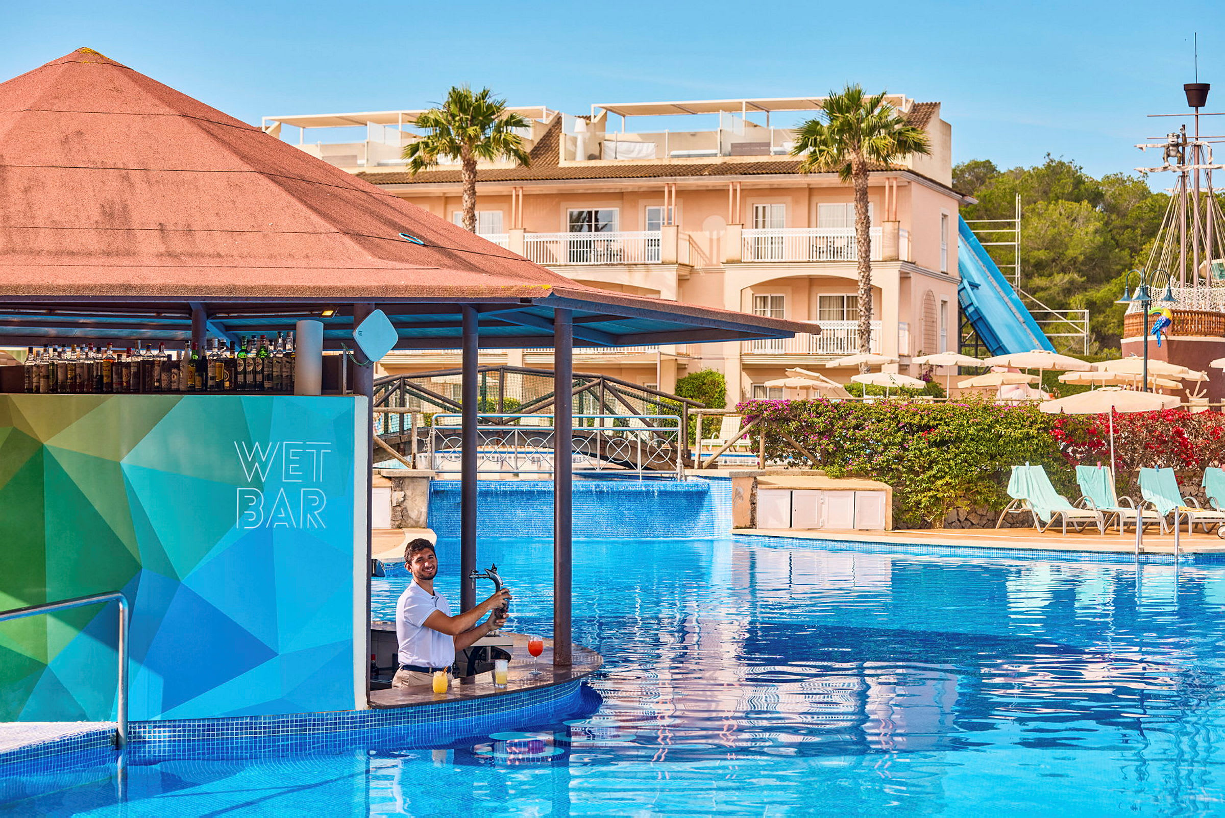 Bar i en swimmingpool med en bartender. I baggrunden ses en bygning, liggestole, parasoller og en vandrutsjebane. (AI-genereret tekst)