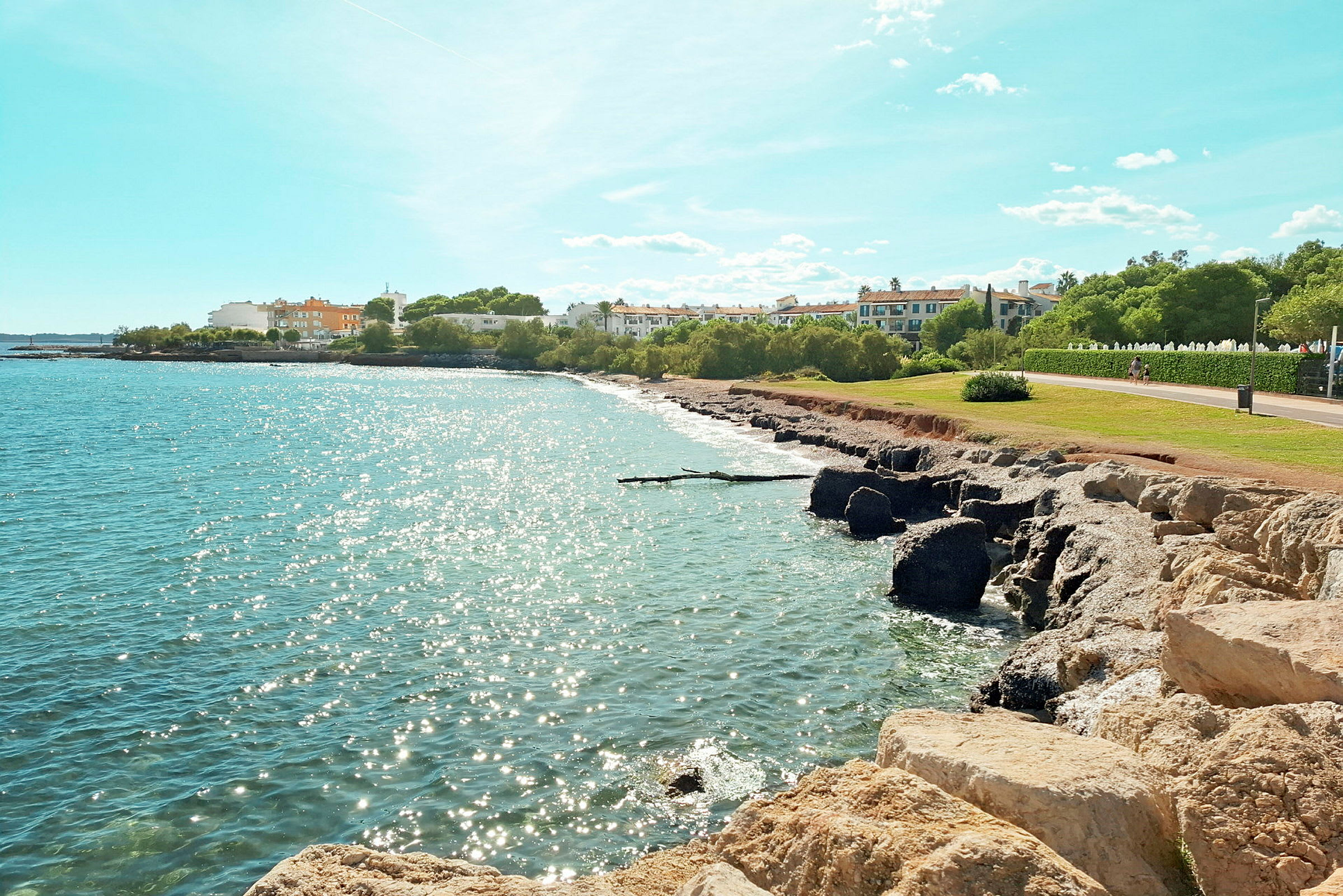En sandstrand med klipper ved en glitrende kyst, med bygninger og grønne områder i baggrunden under en klar blå himmel. (AI-genereret tekst)