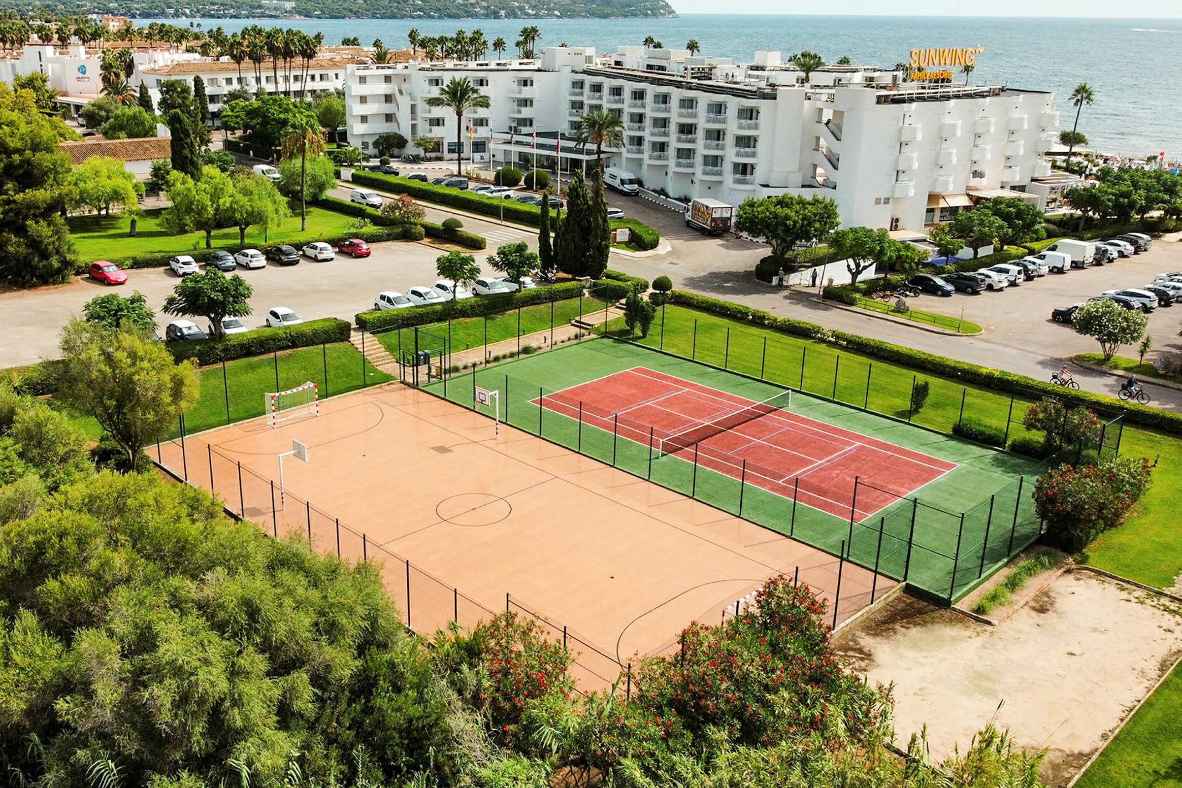 Billede af et tennis- og sportsområde nær havet, med bygninger og parkeringspladser i baggrunden. (AI-genereret tekst)