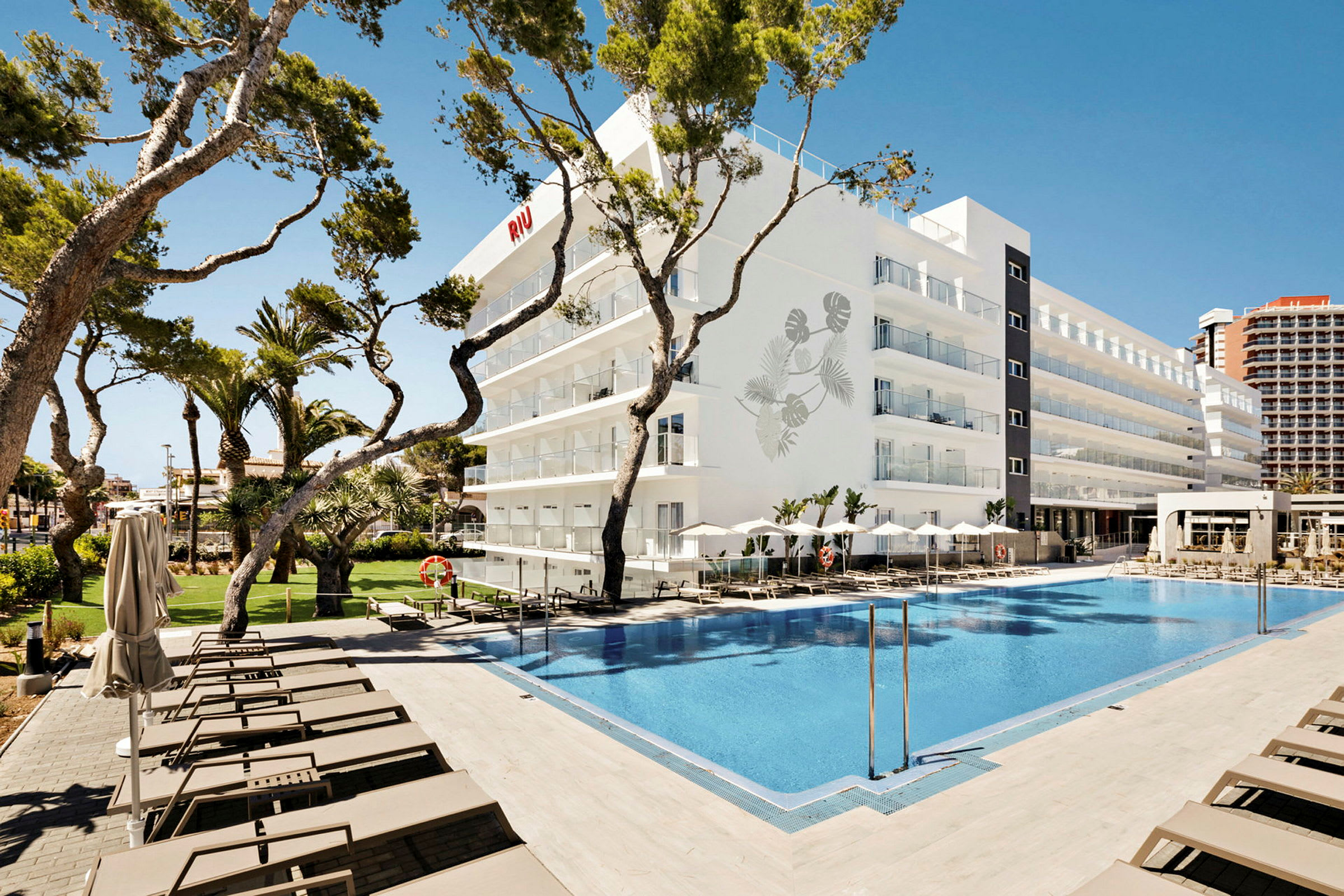 Et hotel med en pool i forgrunden og liggestole omkring, omgivet af træer og blå himmel. (AI-genereret tekst)