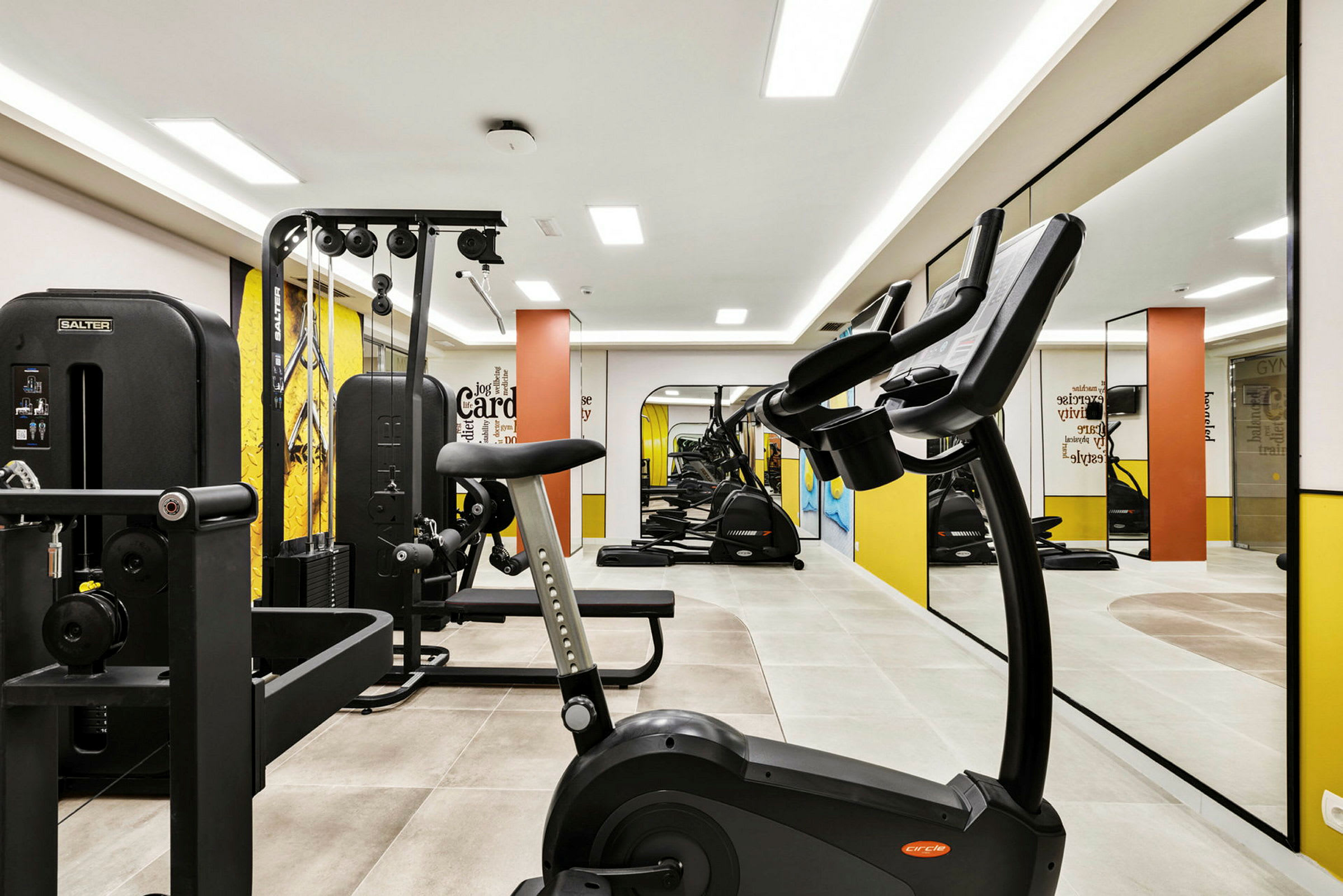 Et fitnesscenter med træningsudstyr såsom motioncykler, styrketræningsmaskiner og spejle på væggen. (AI-genereret tekst)
