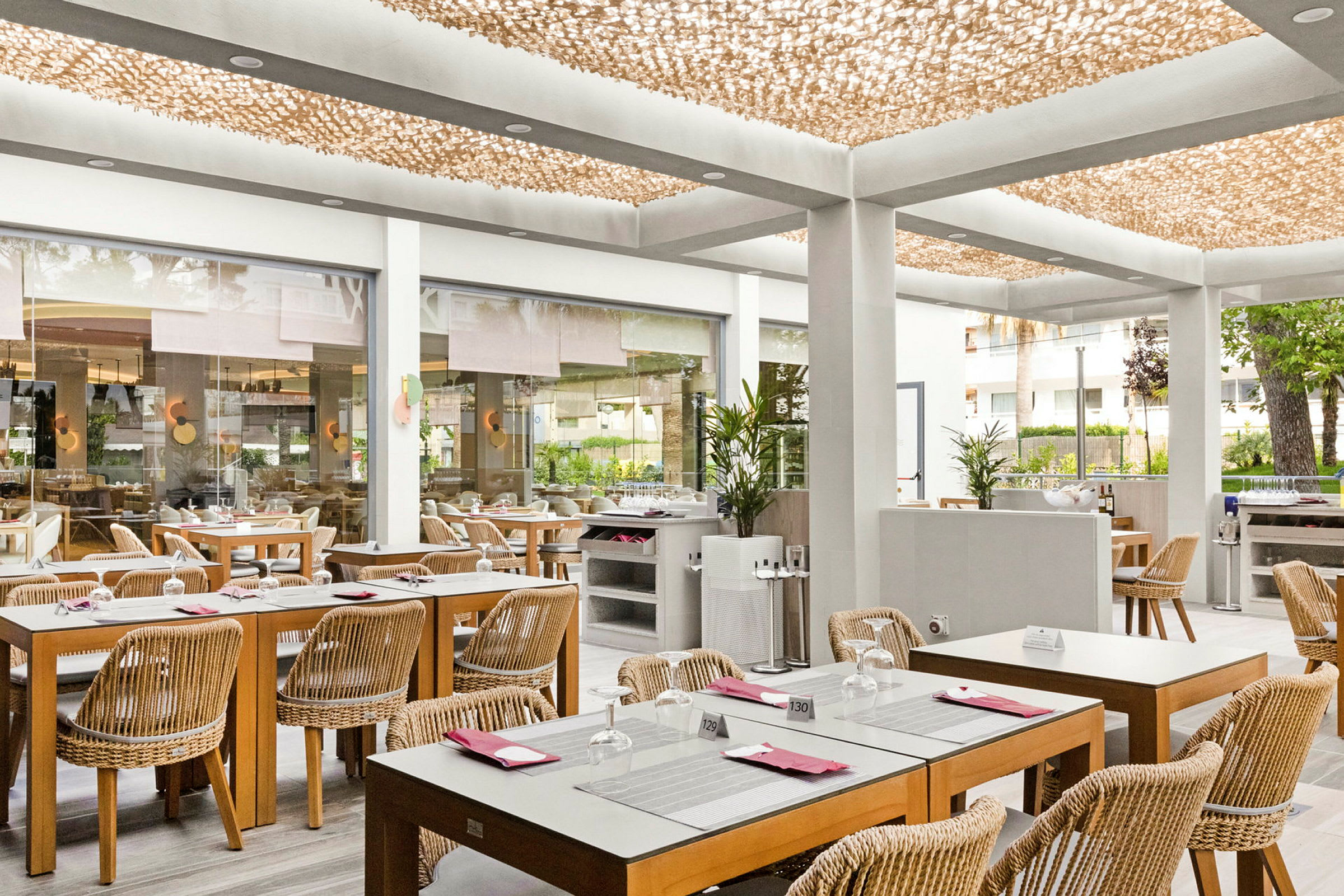En lys restaurant med træborde og rattan-stole, placeret i en moderne indendørs atmosfære med et dekorativt loft og glasvægge, der åbner op mod udendørsområdet. (AI-genereret tekst)