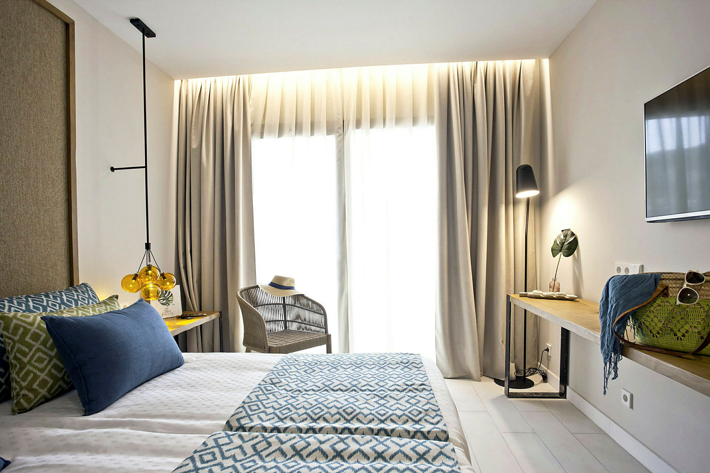 Et lyst hotelværelse med en dobbeltseng, lænestol og et bord med dekorative genstande og lampe. (AI-genereret tekst)