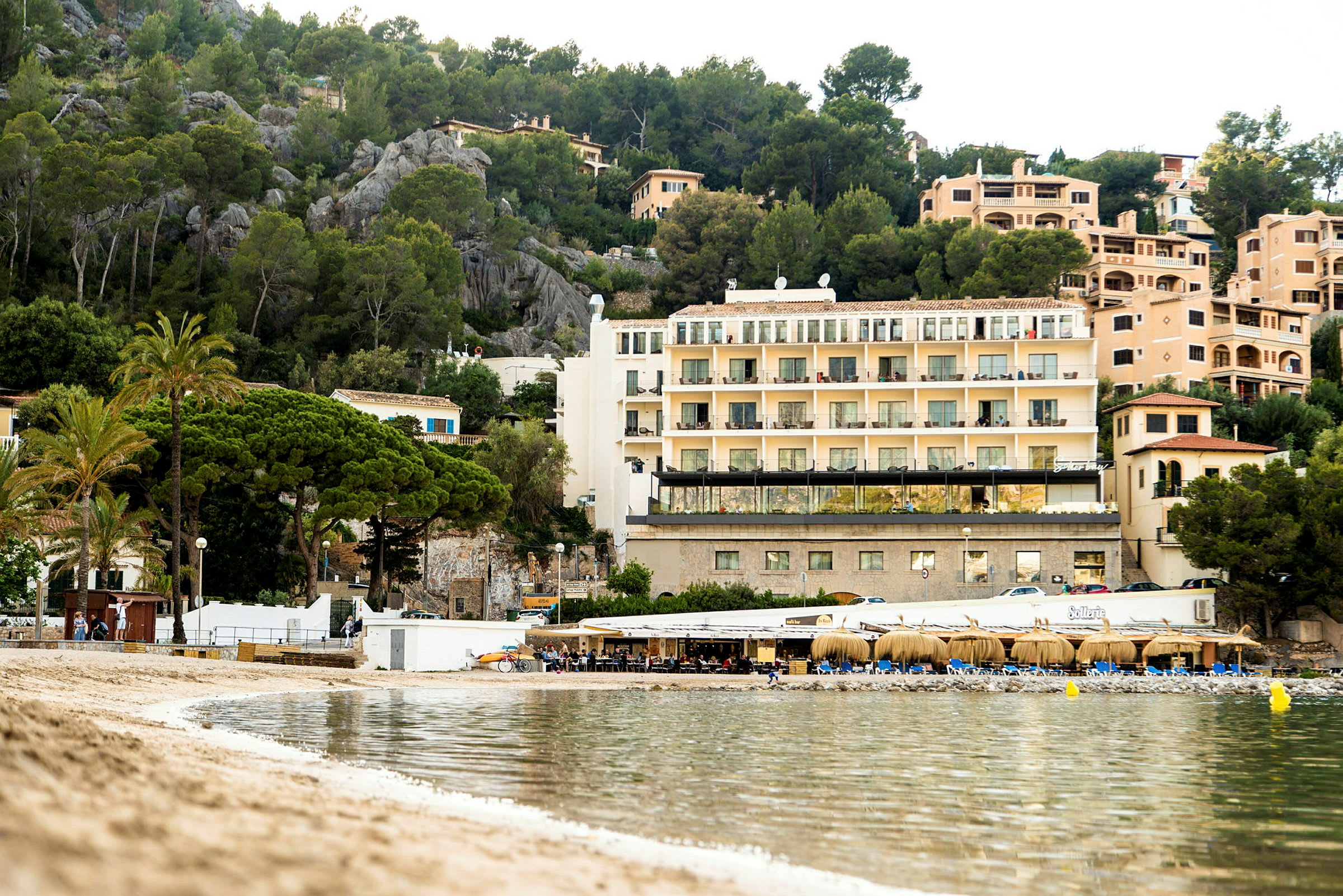 Billede af en strand med klart vand, en lille restaurantbygning og bygninger med flere etager omgivet af grønt i baggrunden. (AI-genereret tekst)