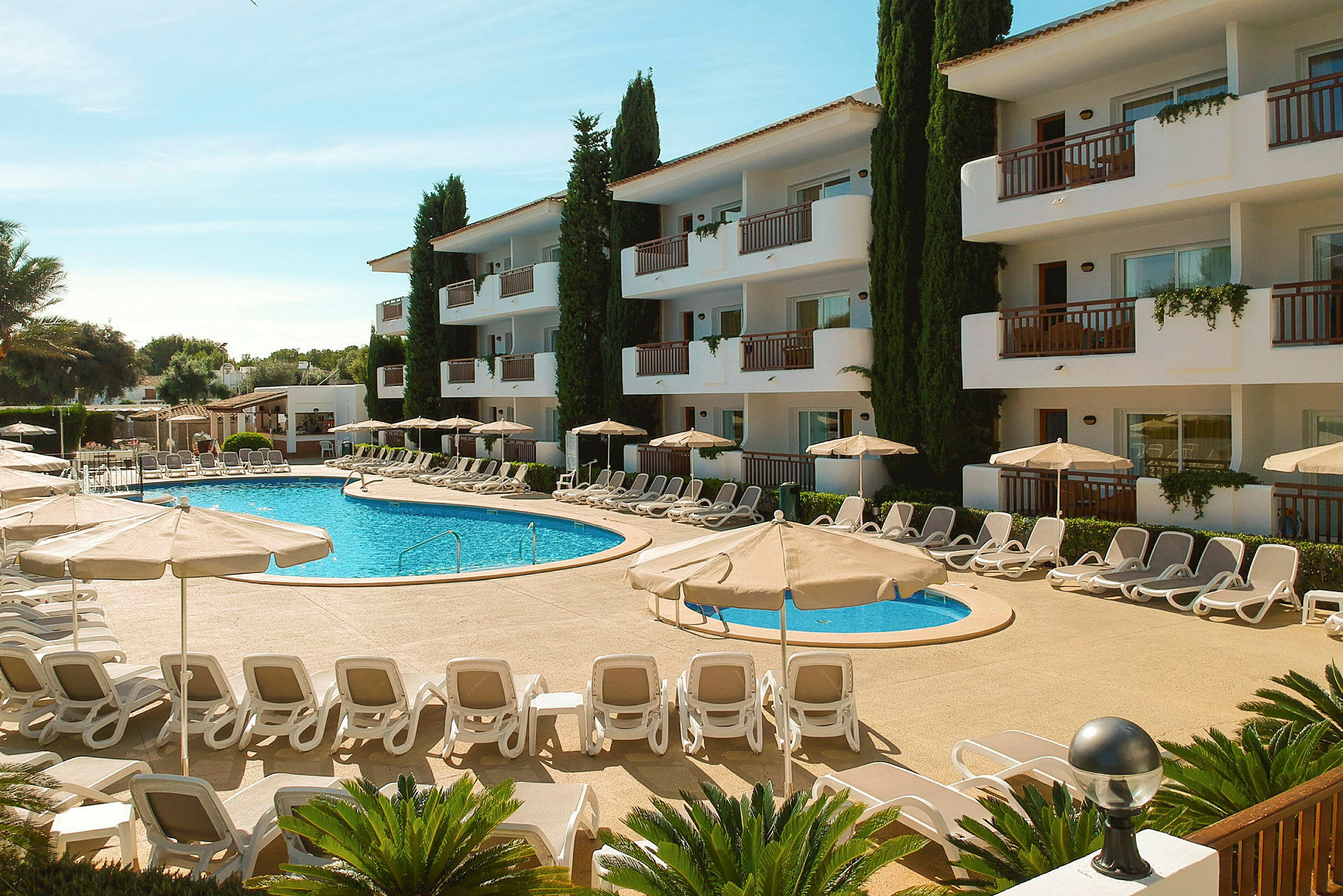 Billede af en hotelpool omgivet af liggestole og parasoller, med en hvid bygning i tre etager i baggrunden. (AI-genereret tekst)