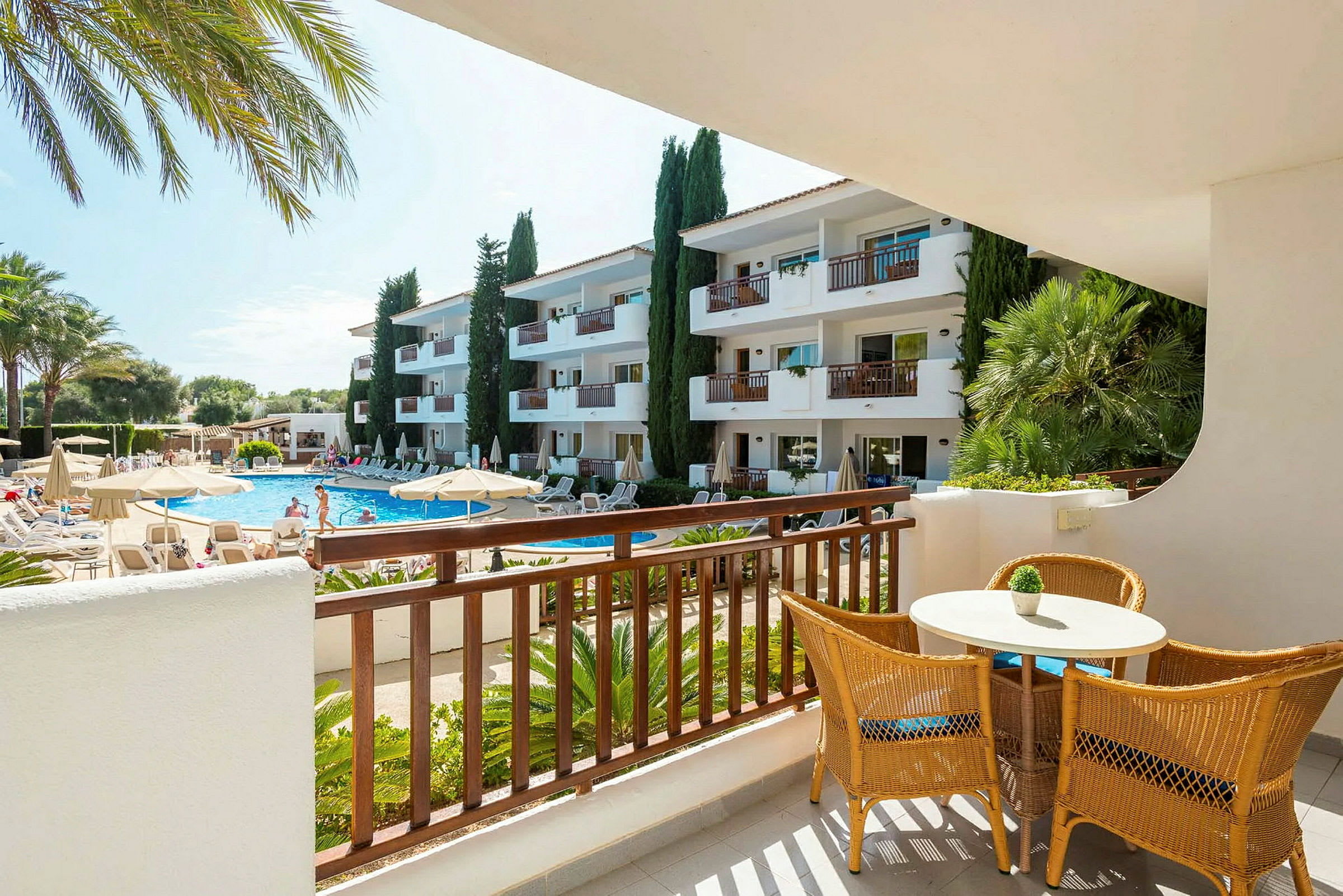 Terrasse med bord og stole med udsigt til poolområde og lejlighedsbygninger. (AI-genereret tekst)