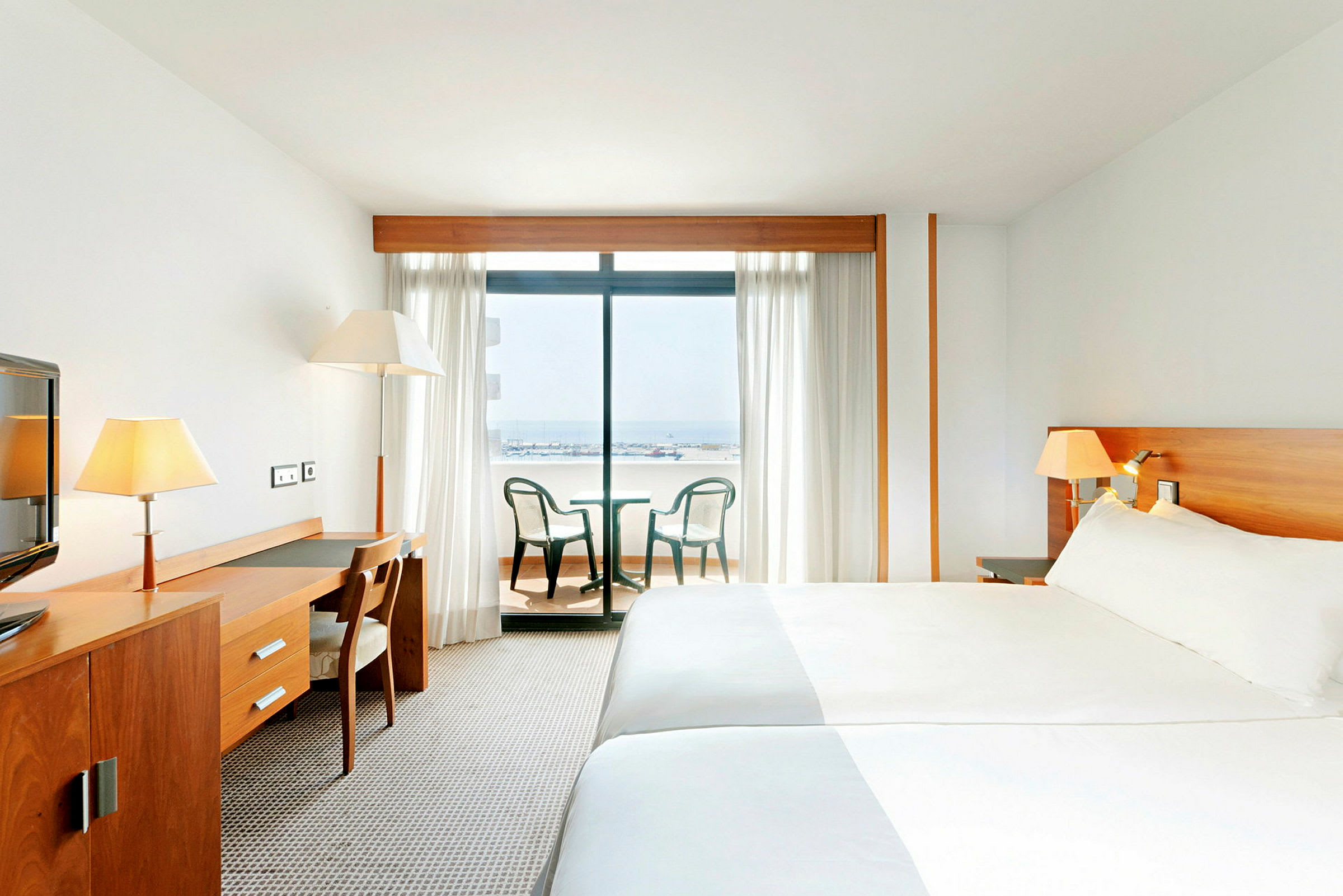 Hotelværelse med en stor seng, et skrivebord med stol og altanmøbler foran en glasdør, der viser en havudsigt. (AI-genereret tekst)