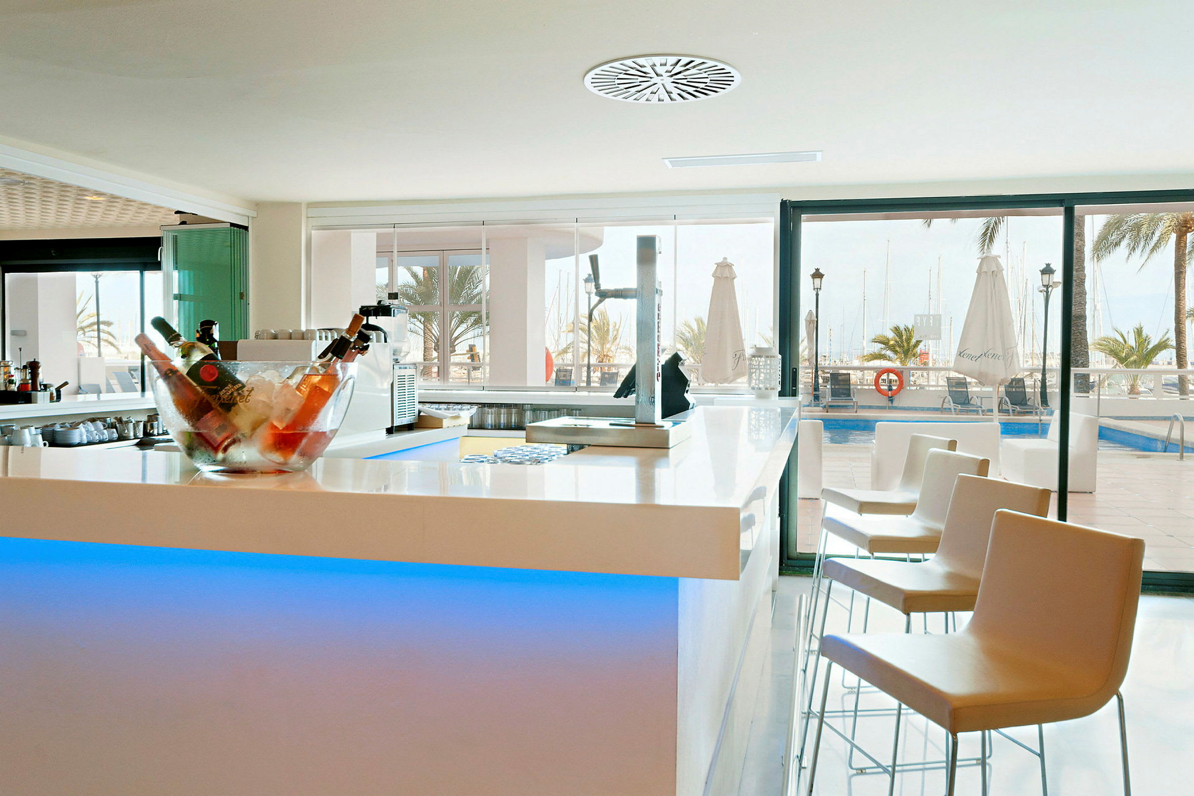 Billede af en moderne bar med lyse møbler, en skål fyldt med flasker på is og udsigt over en pool og marina i baggrunden. (AI-genereret tekst)