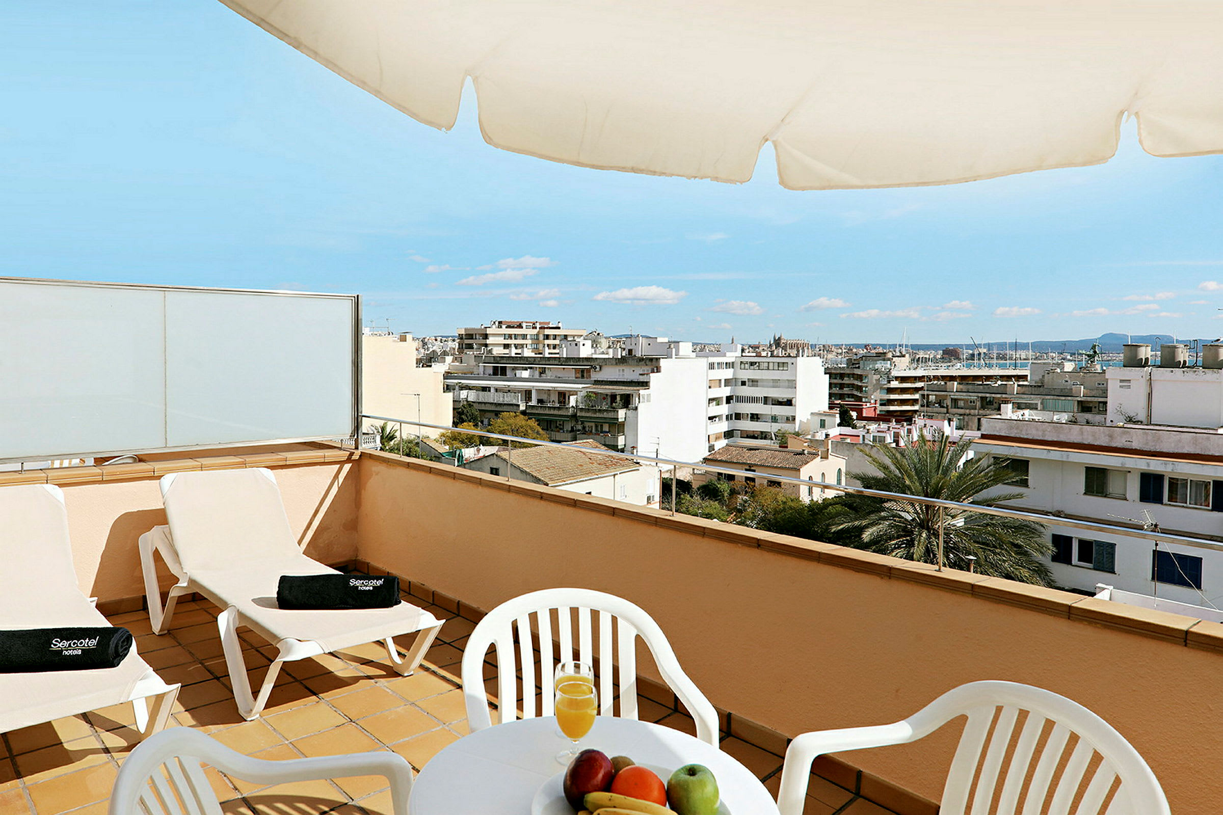 En terrasse med stole, et bord med frugt og juice samt udsigt over en by. (AI-genereret tekst)