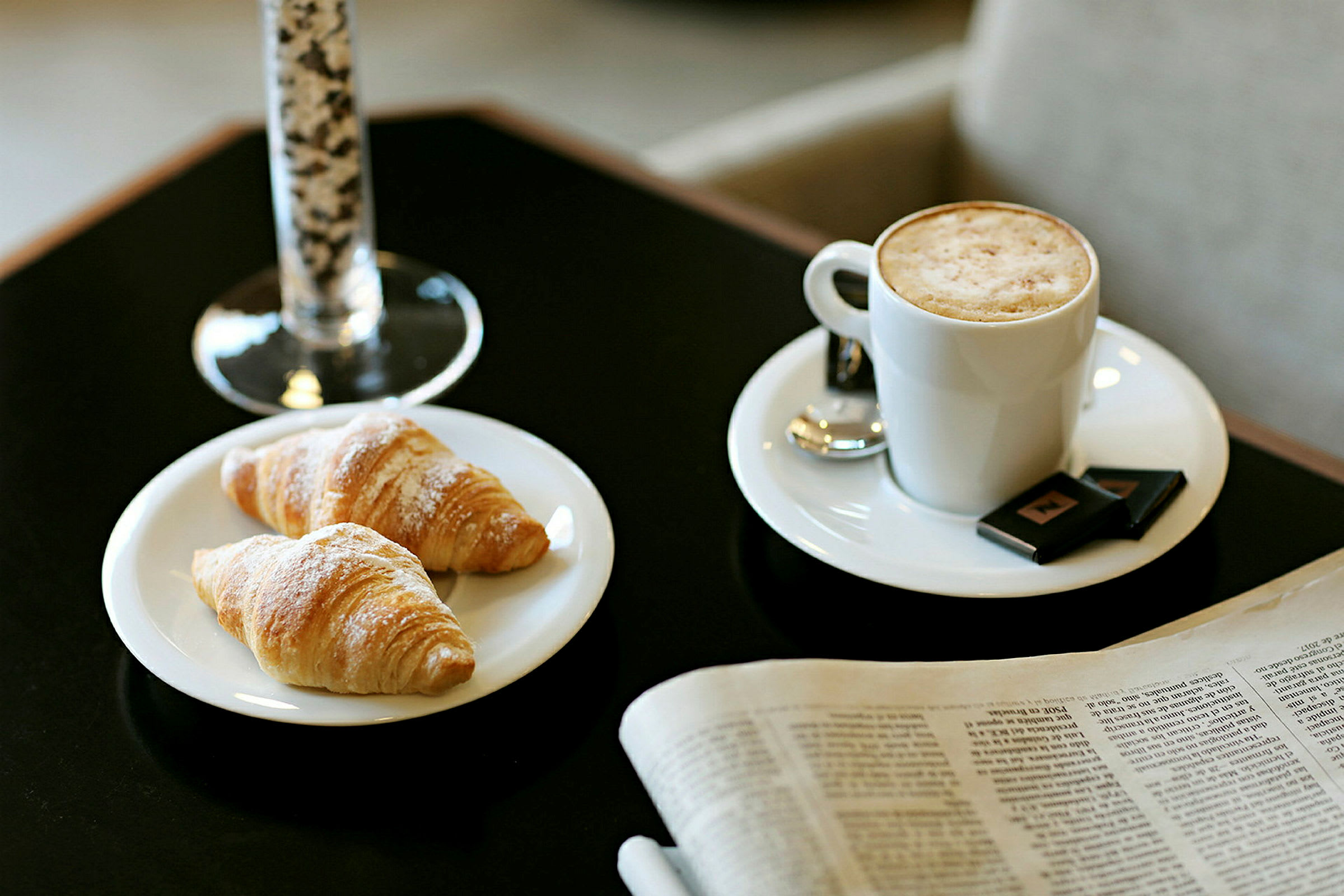 Billedet viser to croissanter på en tallerken, en kop kaffe med mælkeskum og et glas i baggrunden. En avis ligger ved siden af på bordet. (AI-genereret tekst)