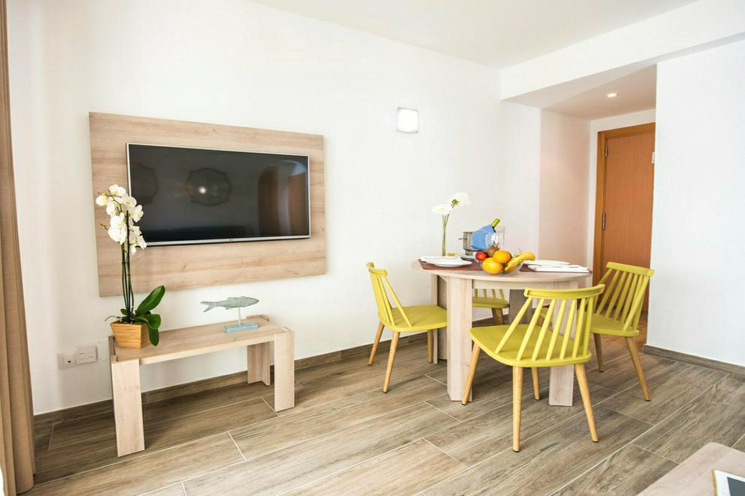 Et værelse med et vægmonteret TV over et bord med en blomsterpotte. På den modsatte side står et rundt spisebord med fire stole, pyntet med frugt og blomster. (AI-genereret tekst)