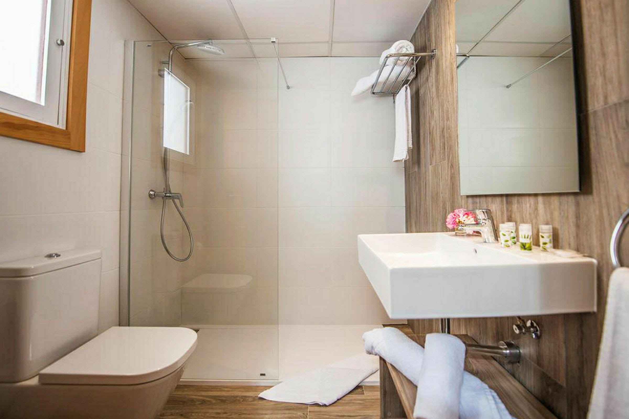 Et badeværelse med bruser, toilet og håndvask. Håndklæder og toiletartikler er placeret på håndvasken. (AI-genereret tekst)