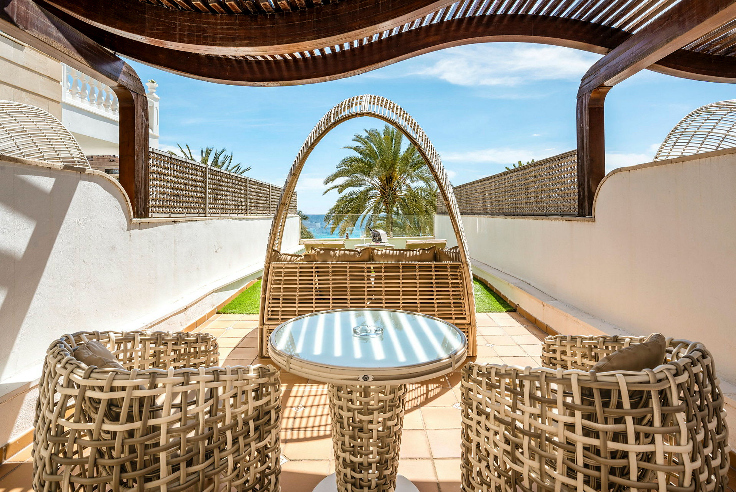 Udendørsmøbler i flettet design på en privat terrasse med udsigt over en palme og havet. (AI-genereret tekst)