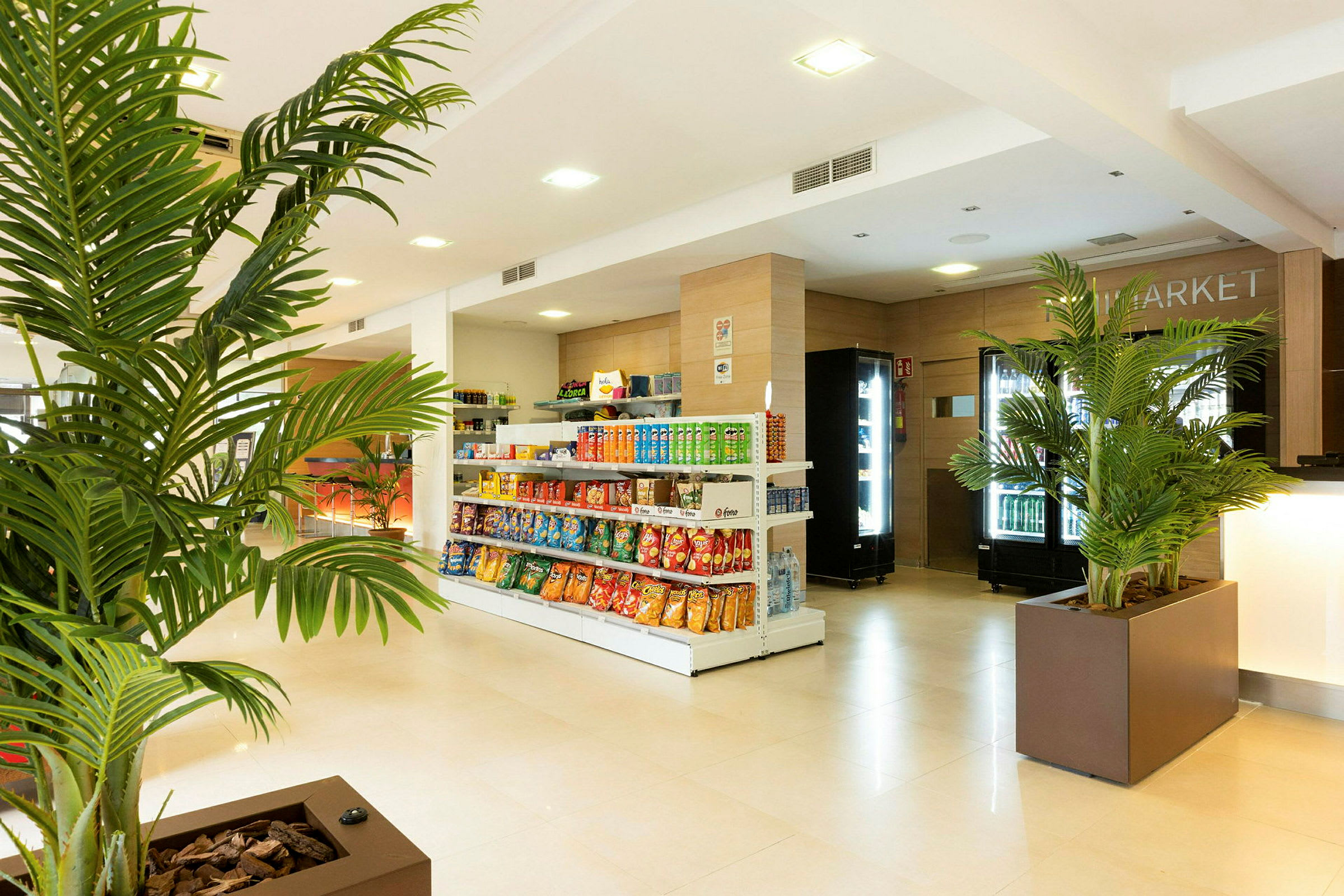 En lys butik med hylder fyldt med snacks og drikkevarer, store grønne planter er placeret som dekoration. (AI-genereret tekst)