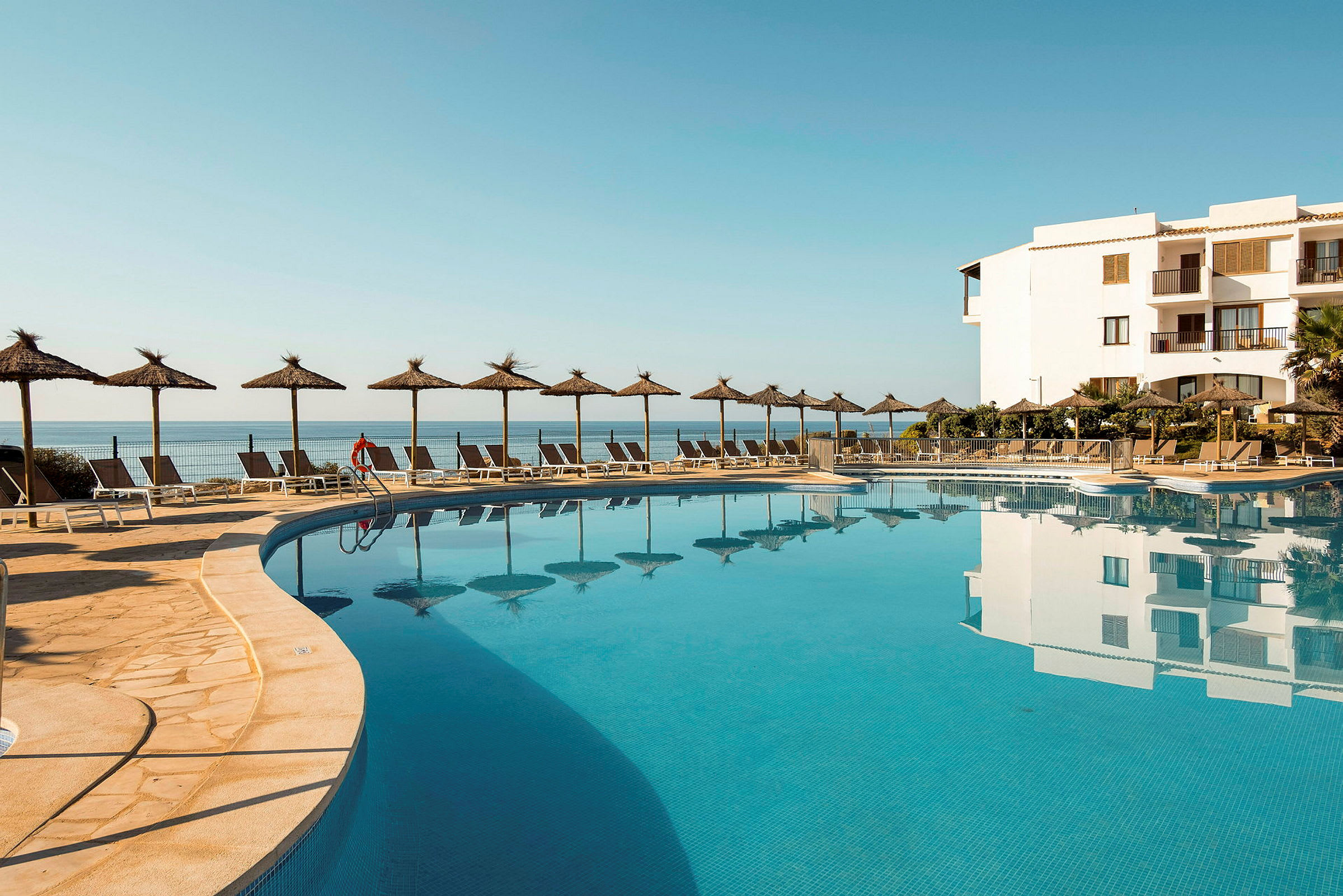 En udendørs pool ved et hotel med parasoller og liggestole, havet i baggrunden. (AI-genereret tekst)