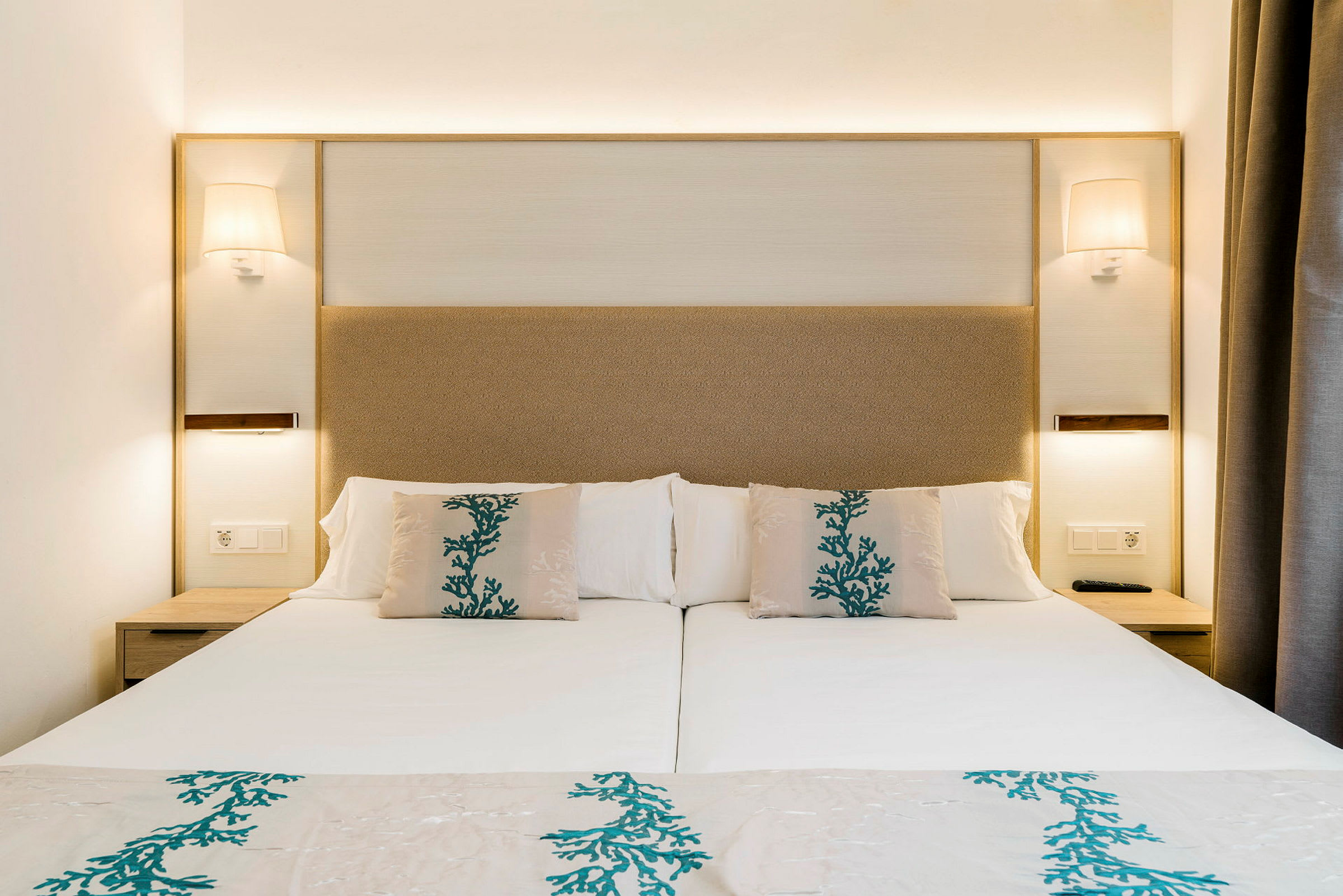 En hotelseng med hvidt sengetøj og puder dekoreret med blågrønt mønster. (AI-genereret tekst)