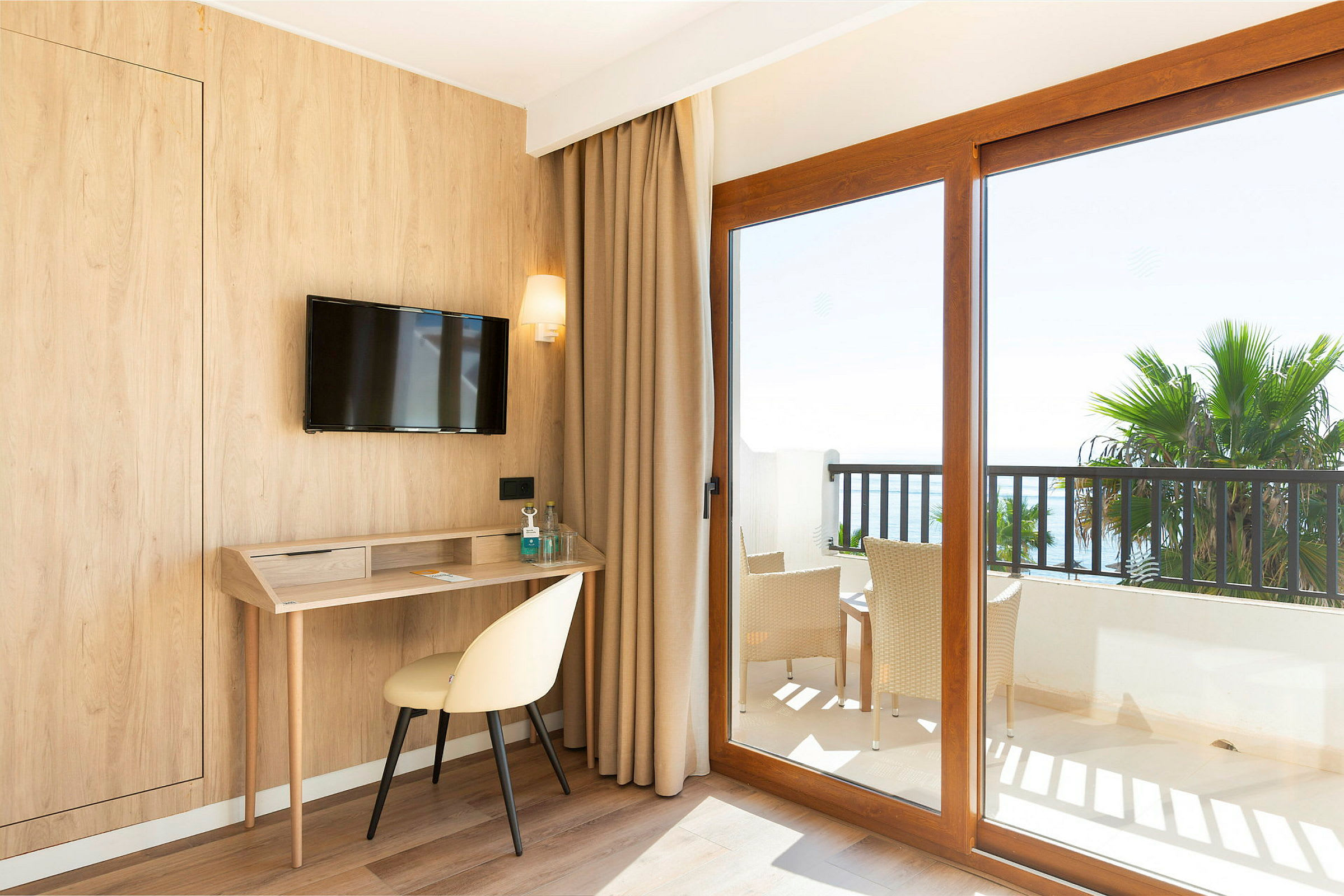 Hotelværelse med træpanel, skrivebord, stol og en TV på væggen. Store vinduer med udsigt til en balkon med møbler og palmer. (AI-genereret tekst)