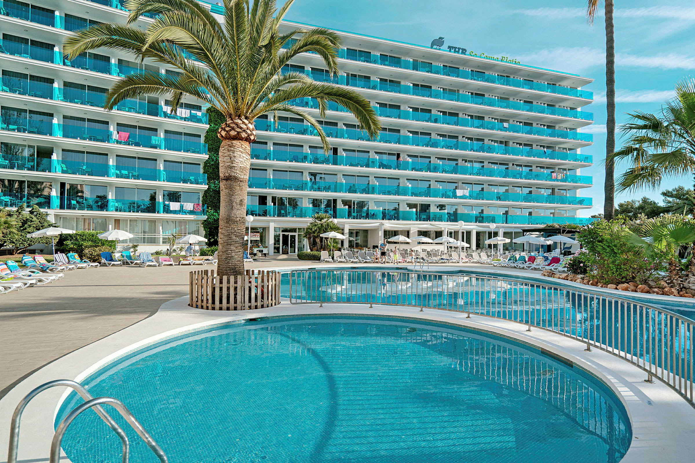 Billede af et hotel med en pool, omgivet af liggestole og parasoller, med palmer og grønne planter. (AI-genereret tekst)