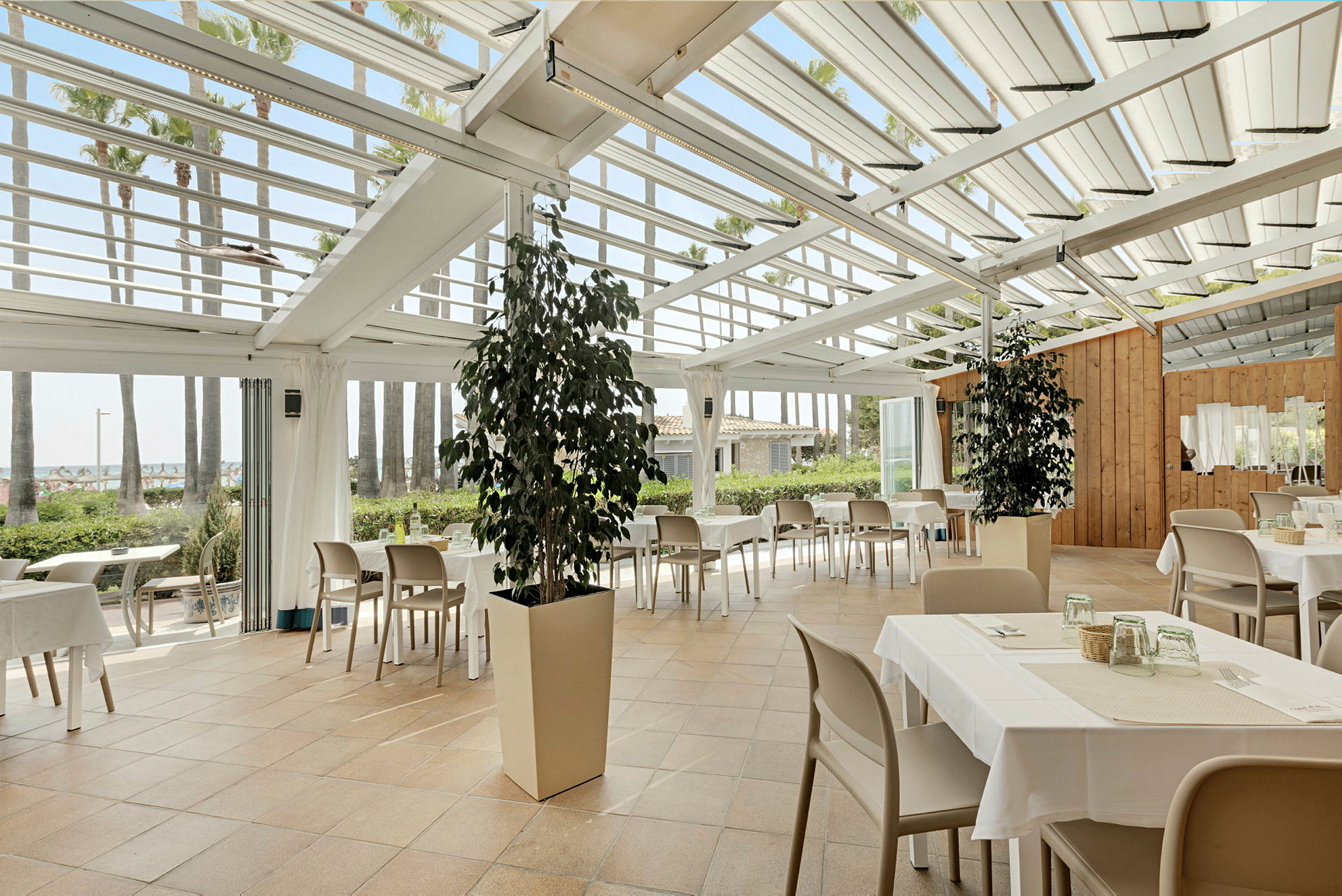 Restaurant med borde og stole under et glastag, omgivet af grønne planter og palmer. (AI-genereret tekst)