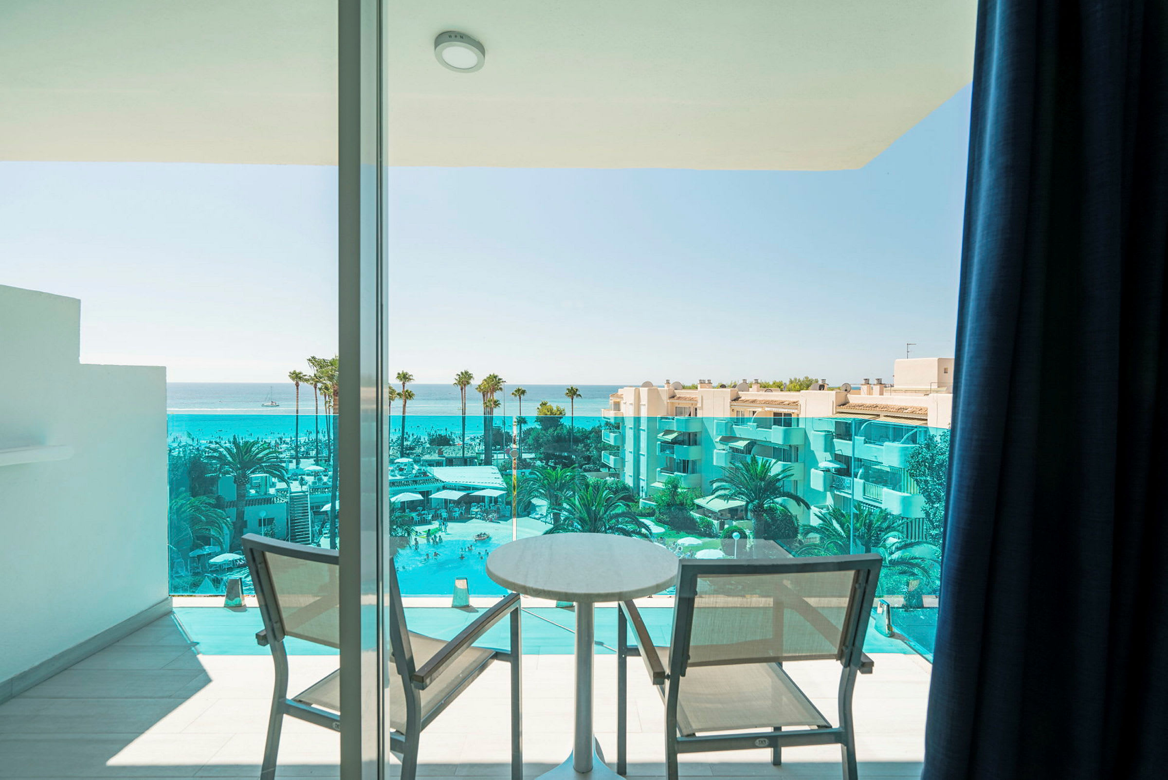 Udsigt fra en altan med to stole og et bord mod havet og en hotelpool. (AI-genereret tekst)