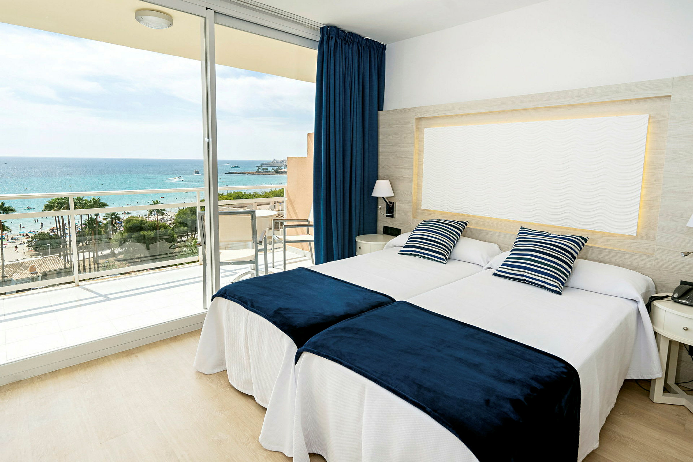 Et hotelværelse med to senge, blåhvide pyntepuder og blå sengetæpper samt en balkon med udsigt til havet. (AI-genereret tekst)