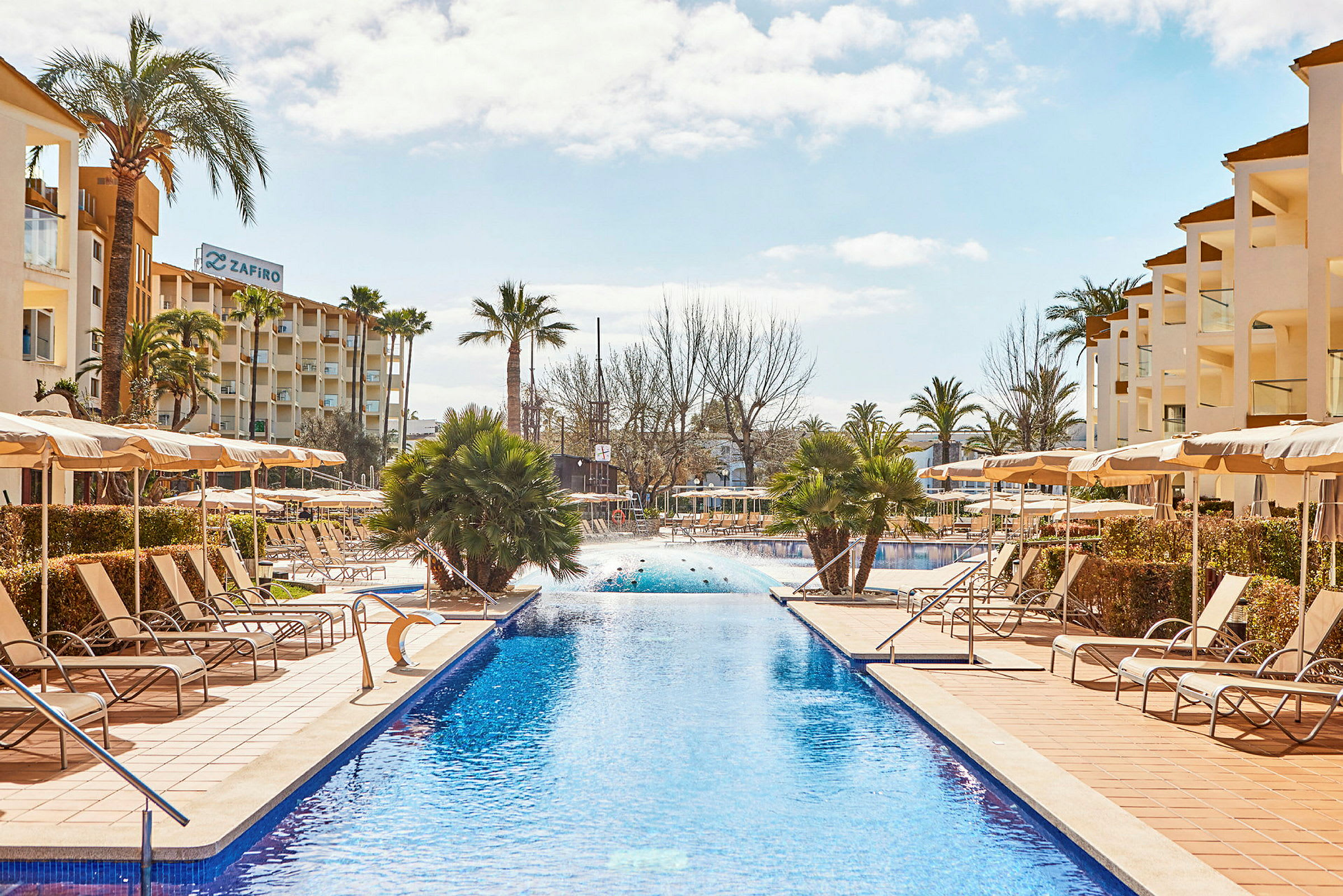 Billede af en lang swimmingpool omgivet af liggestole og parasoller, med hotelbygninger i baggrunden. (AI-genereret tekst)