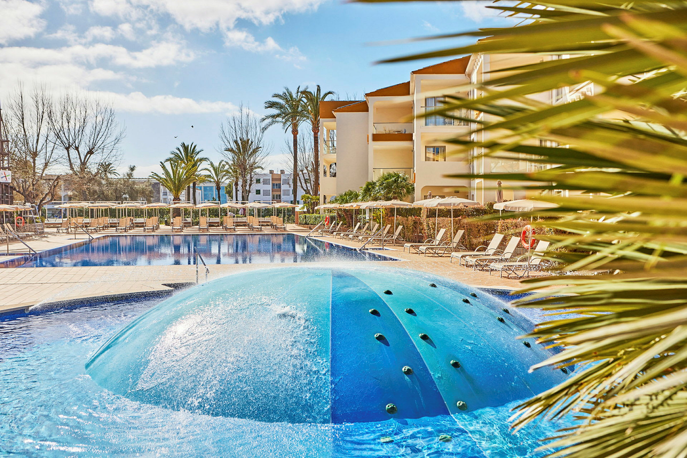 Billede af en pool med en bølgeformet springvand i forgrunden, omgivet af liggestole og et hotel i baggrunden. (AI-genereret tekst)