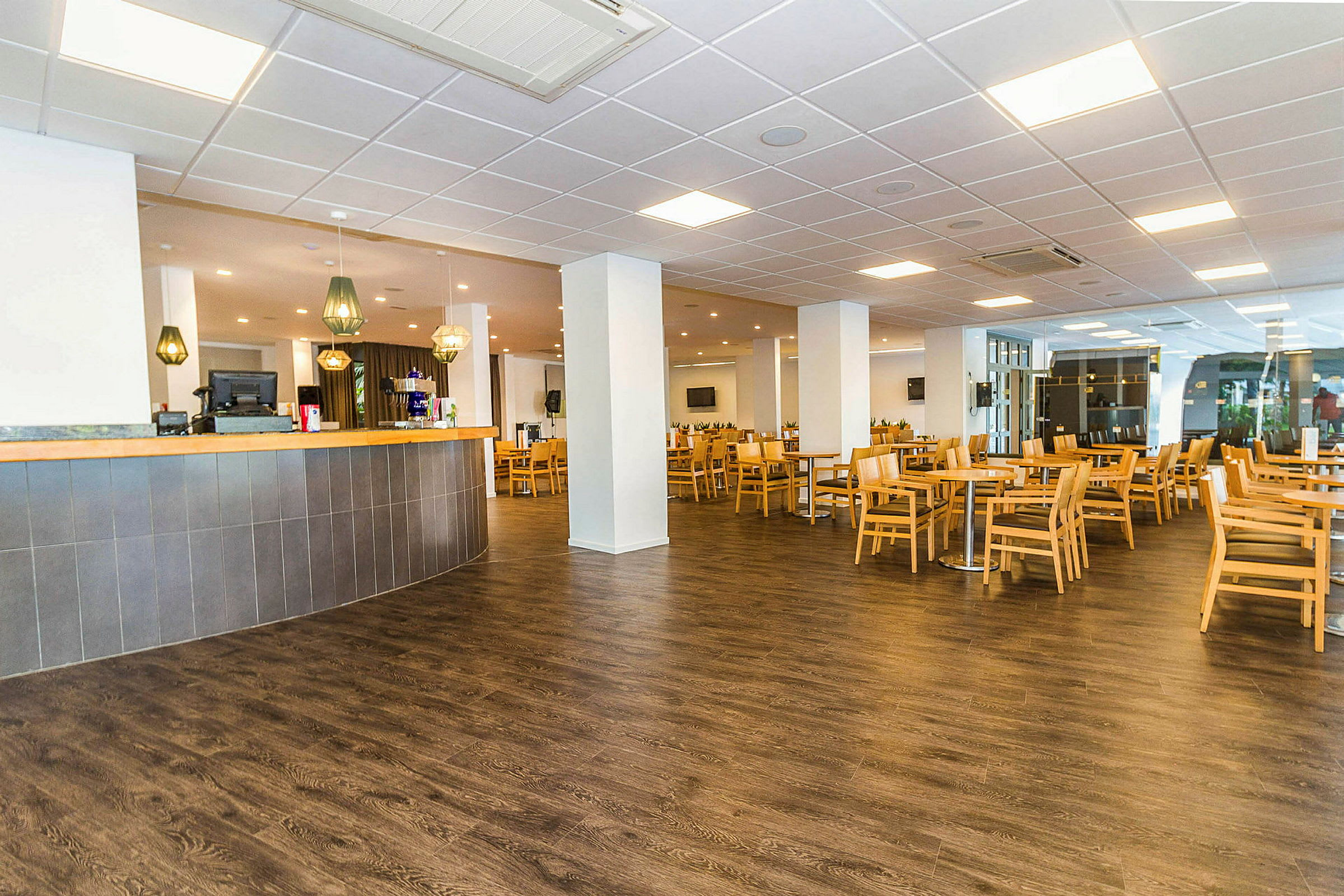Et rummeligt café- eller restaurantområde med lyse borde og træstole placeret på et mørkt gulv. I forgrunden er der en bar med hængende lamper over. (AI-genereret tekst)
