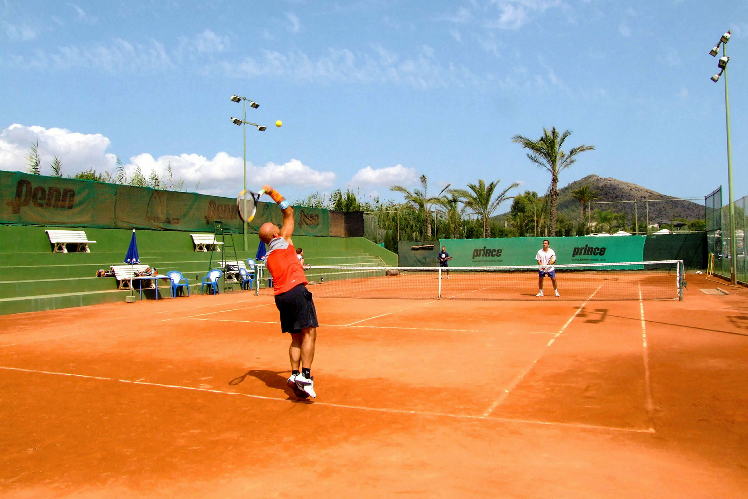 To personer spiller tennis på en grusbane udendørs på en solrig dag. (AI-genereret tekst)