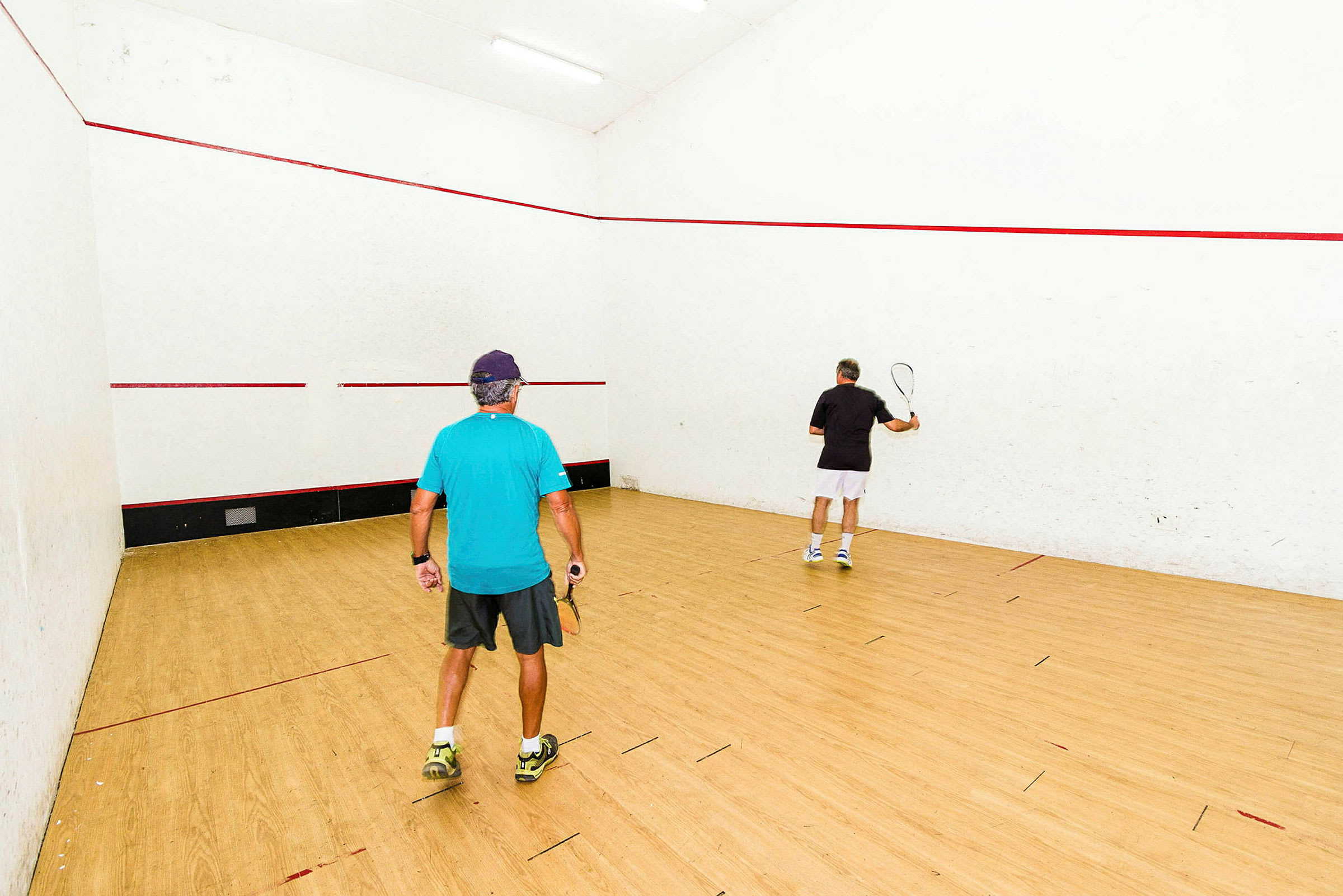 To personer spiller squash på en indendørsbane med lyse vægge og trægulv. (AI-genereret tekst)