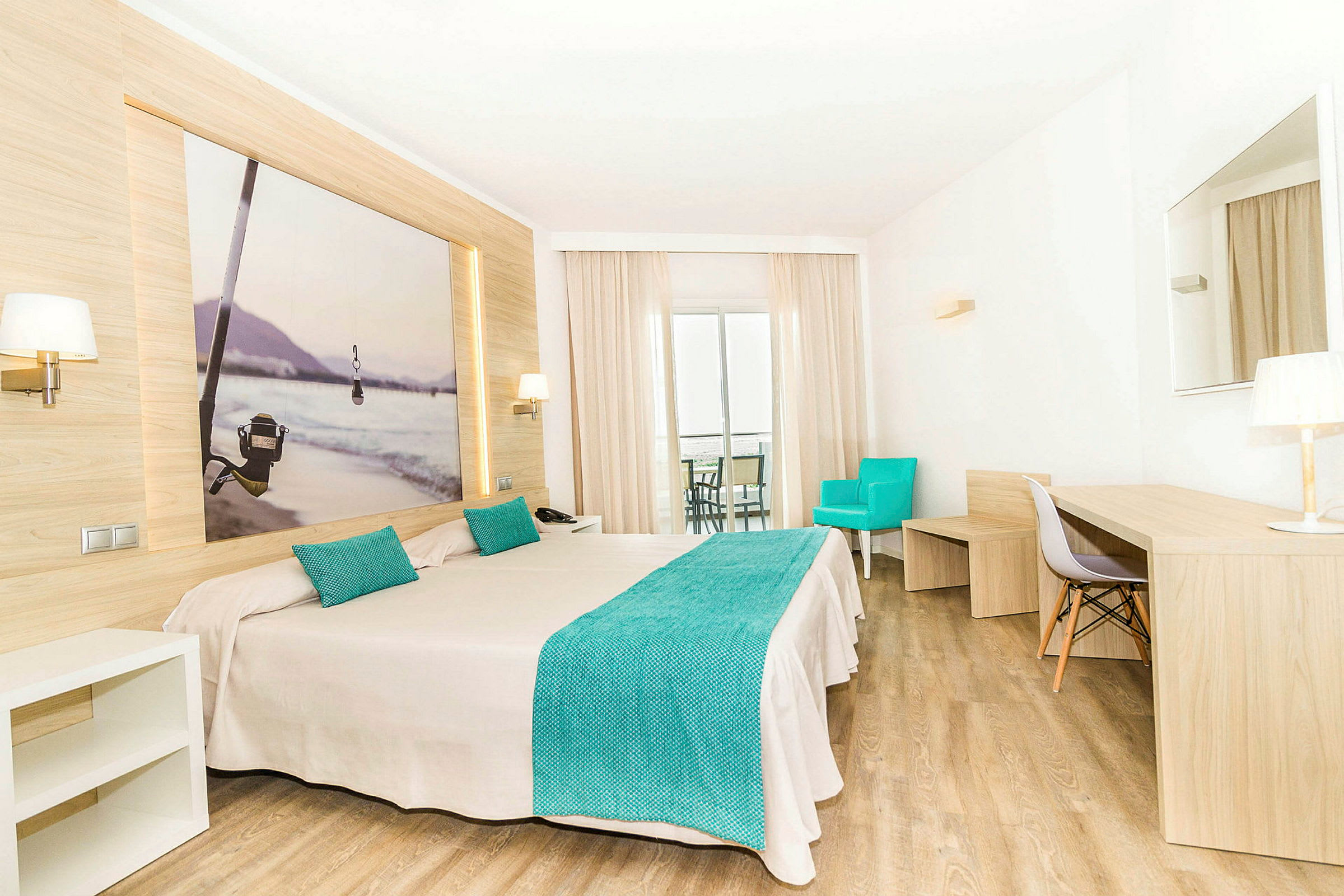 Et hotelværelse med en dobbeltseng, turkise detaljer og moderne trædesign. Bag sengen hænger et billede med et fiske-tema. Værelset har et skrivebord, en turkis lænestol og en altan. (AI-genereret tekst)
