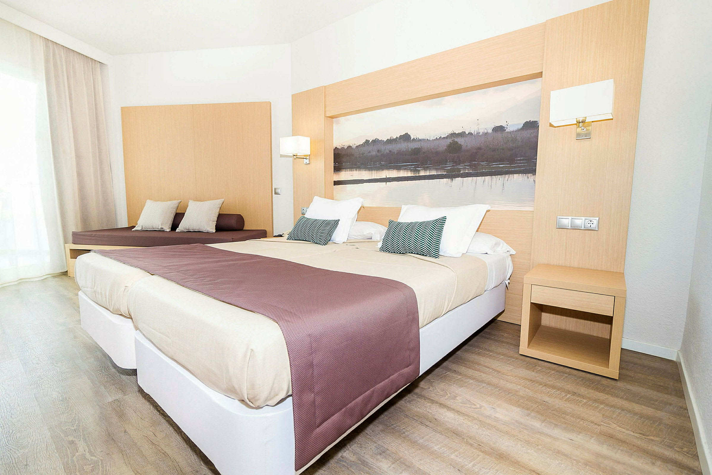 Et lyst hotelværelse med to senge og en siddeplads. Sengene er redt med beige og lilla sengetæpper, og et stort billede pryder væggen bag sengene. (AI-genereret tekst)