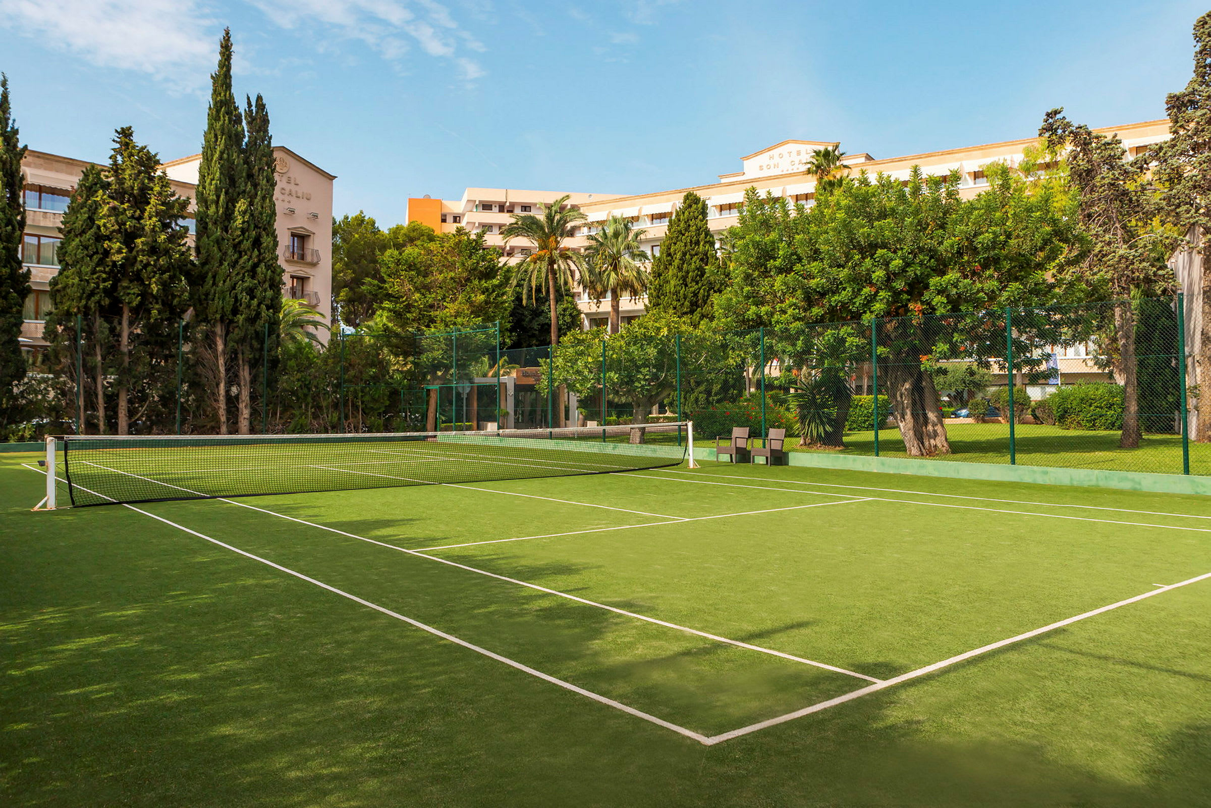En tennisbane med grønt underlag og hvide linjer, omgivet af træer og bygninger. (AI-genereret tekst)