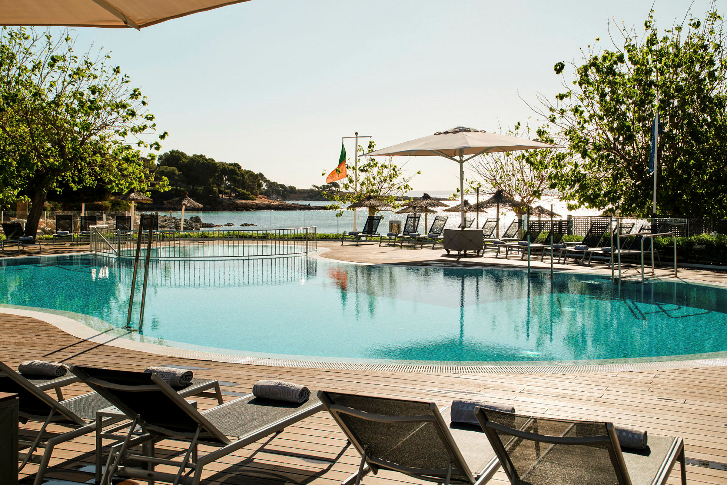 Pool med liggestole, træer og hav i baggrunden. (AI-genereret tekst)