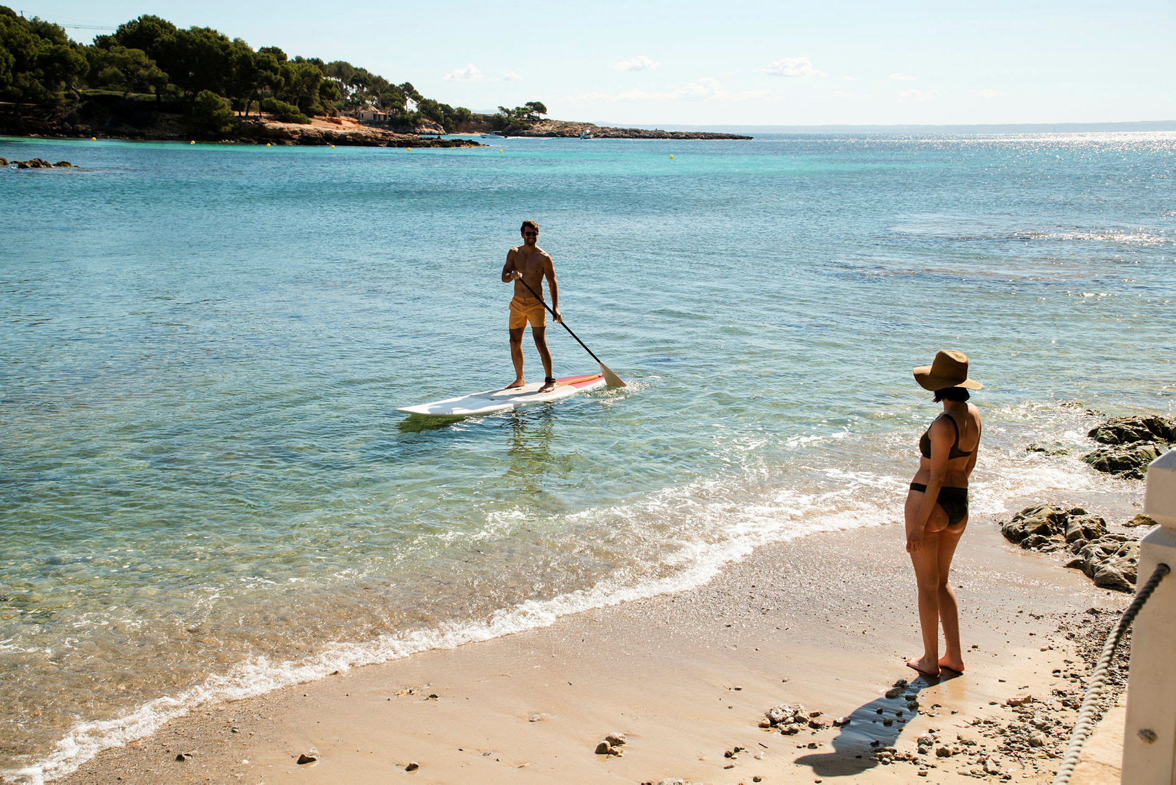 En person står på et paddleboard i havet nær stranden, mens en anden person står på sandstranden og kigger mod vandet. (AI-genereret tekst)