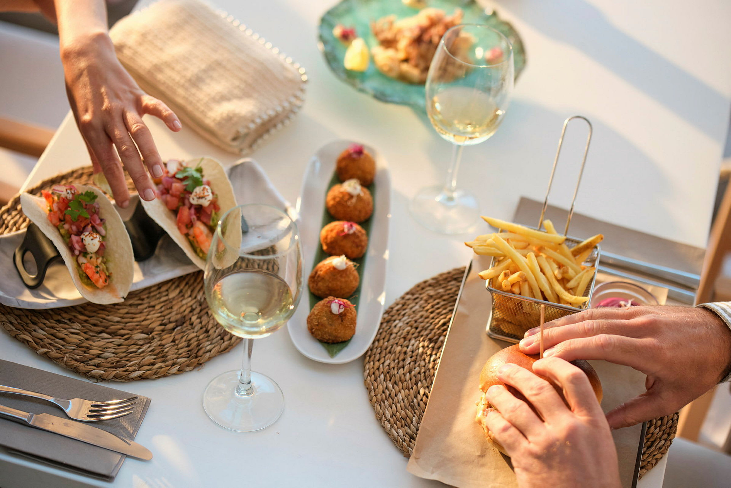 Nærbillede af en bordscene med mad og drikke, herunder tacos, croquetas, pommes frites, en burger og vinglas. (AI-genereret tekst)