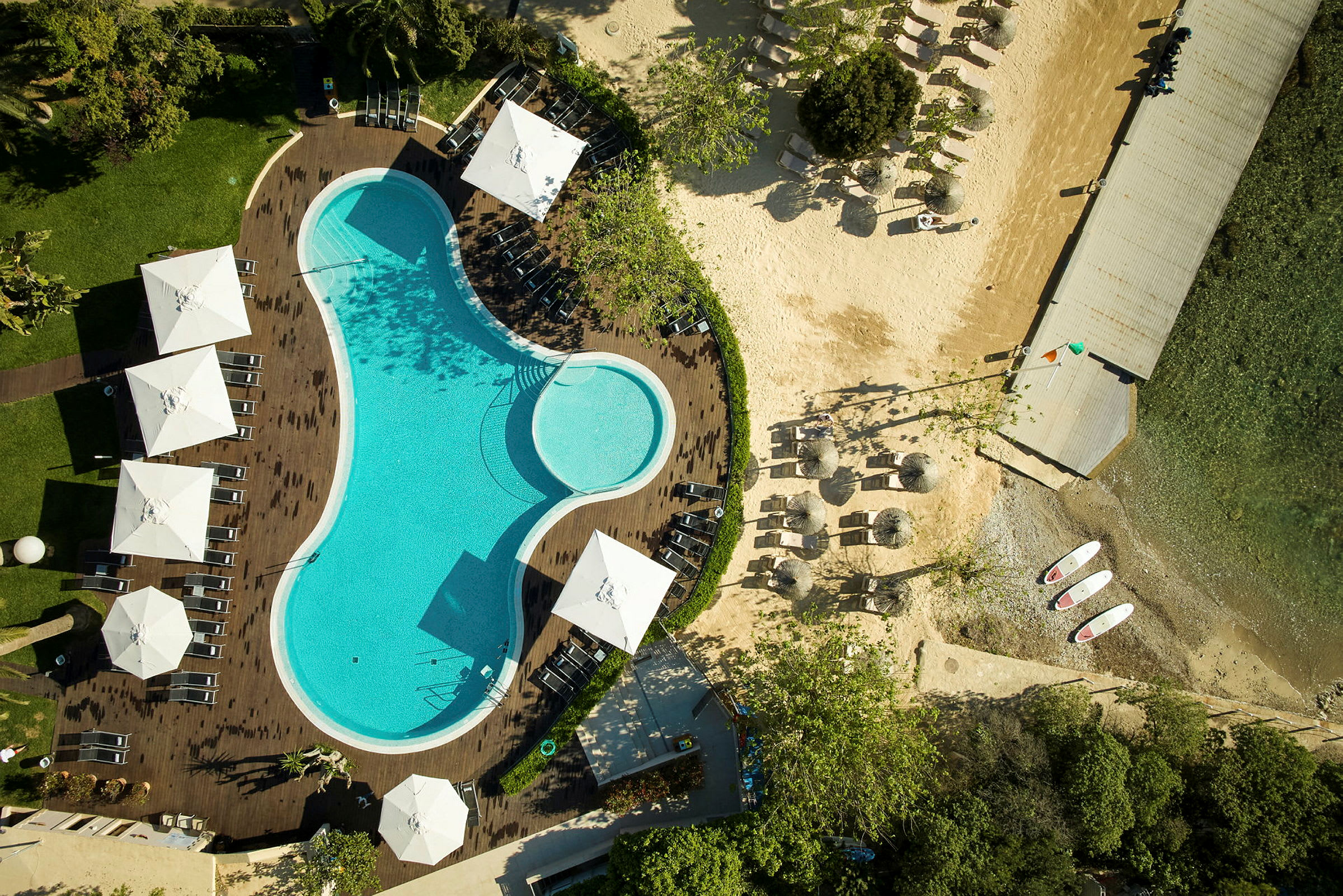 Luftfoto af en pool omgivet af liggestole og parasoller, med en tilstødende strand og bro. (AI-genereret tekst)