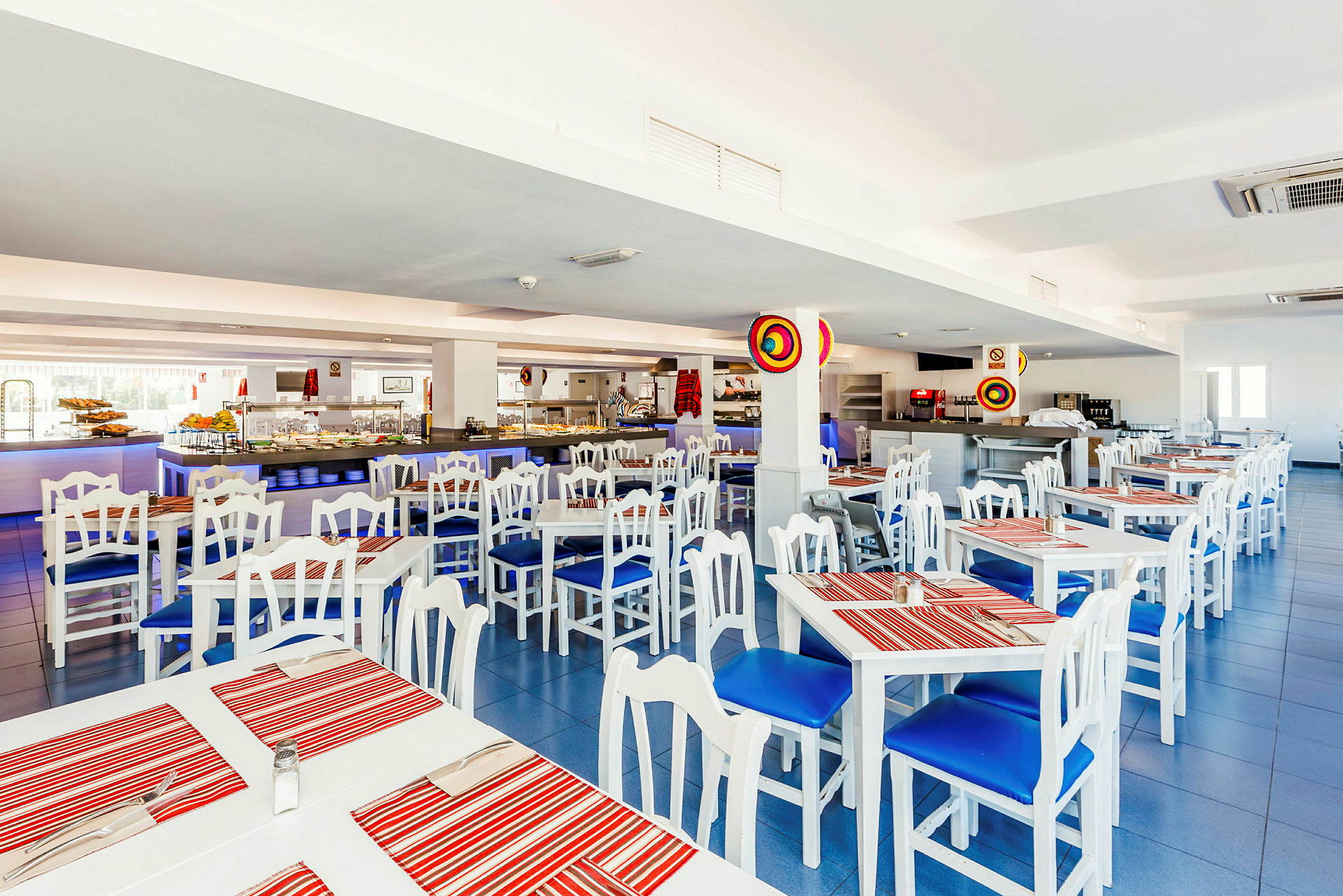 Interiør af en restaurant med hvide borde og stole med blå hynder samt røde stribede bordskånere. (AI-genereret tekst)