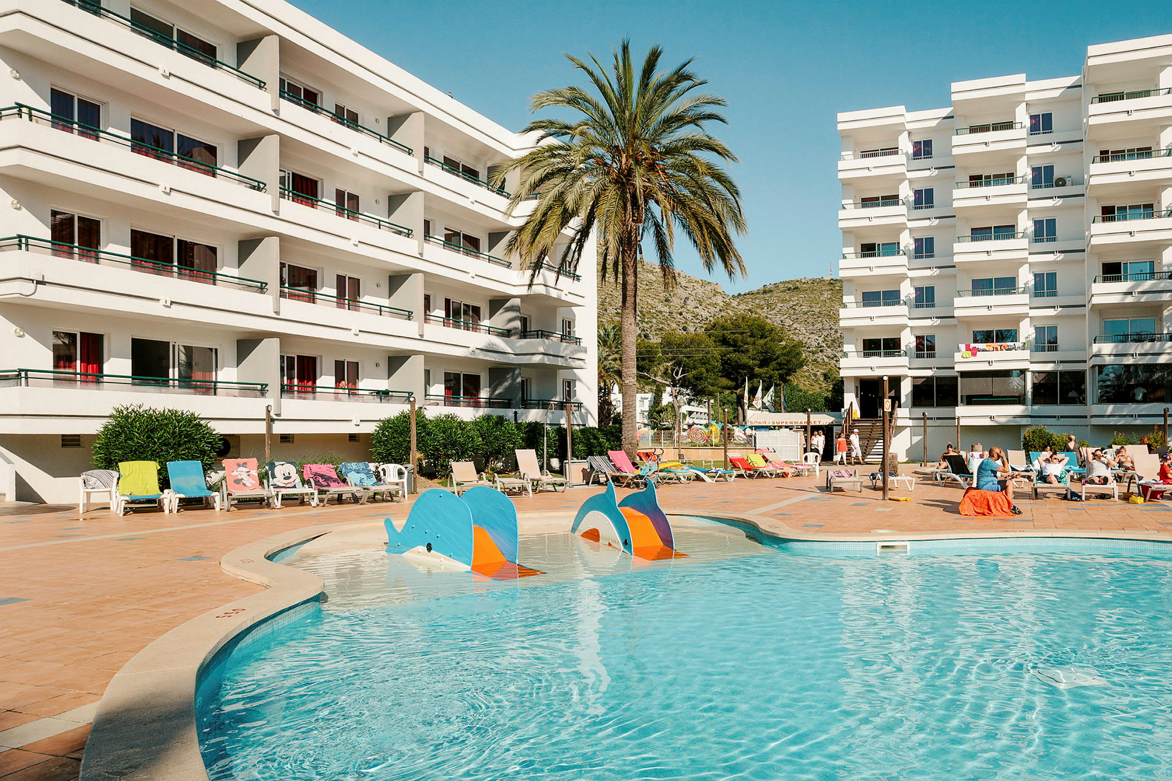 Hotelbygning med altaner ved en udendørs pool og liggestole. (AI-genereret tekst)