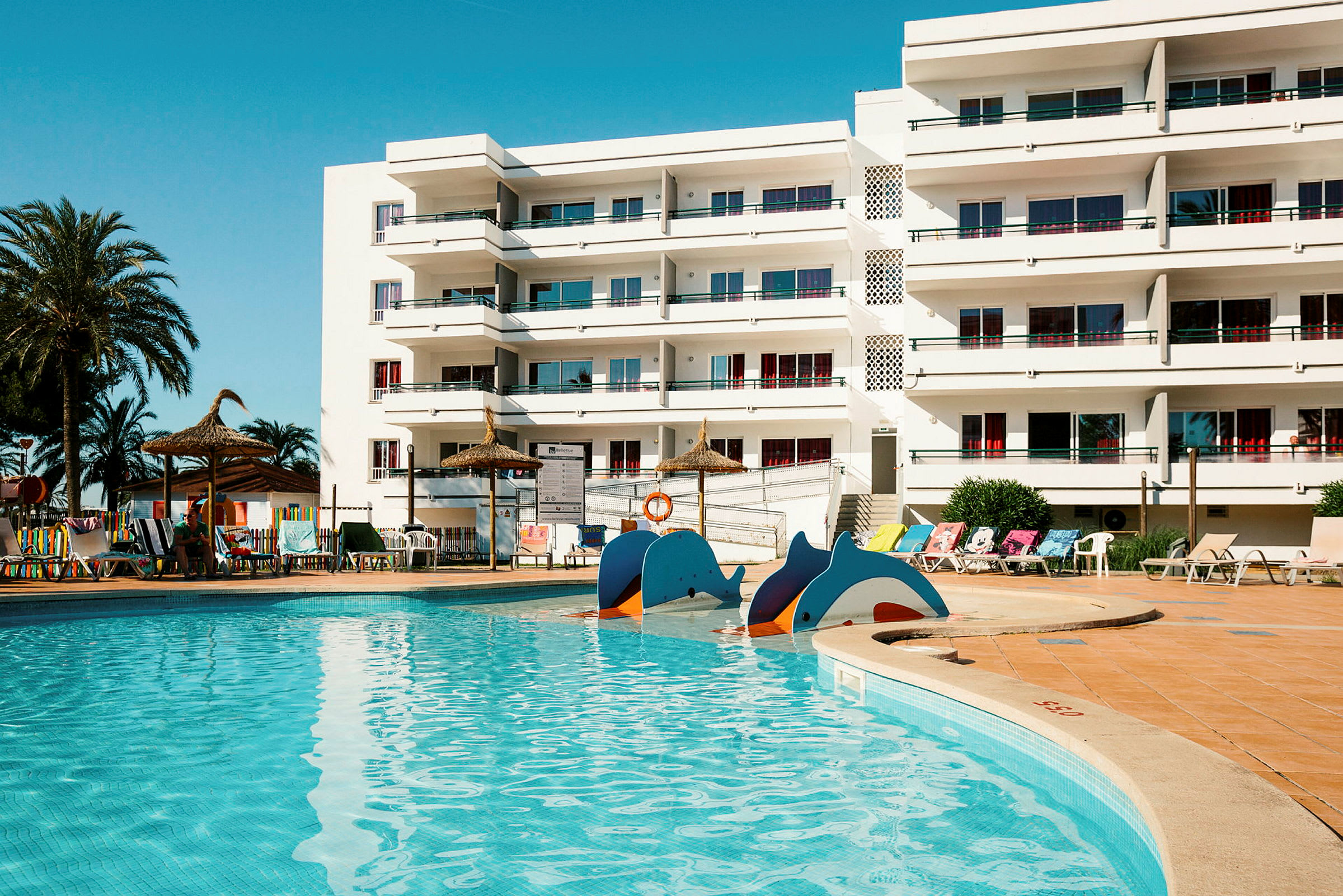 En swimmingpool foran en hvid bygning med altaner. (AI-genereret tekst)